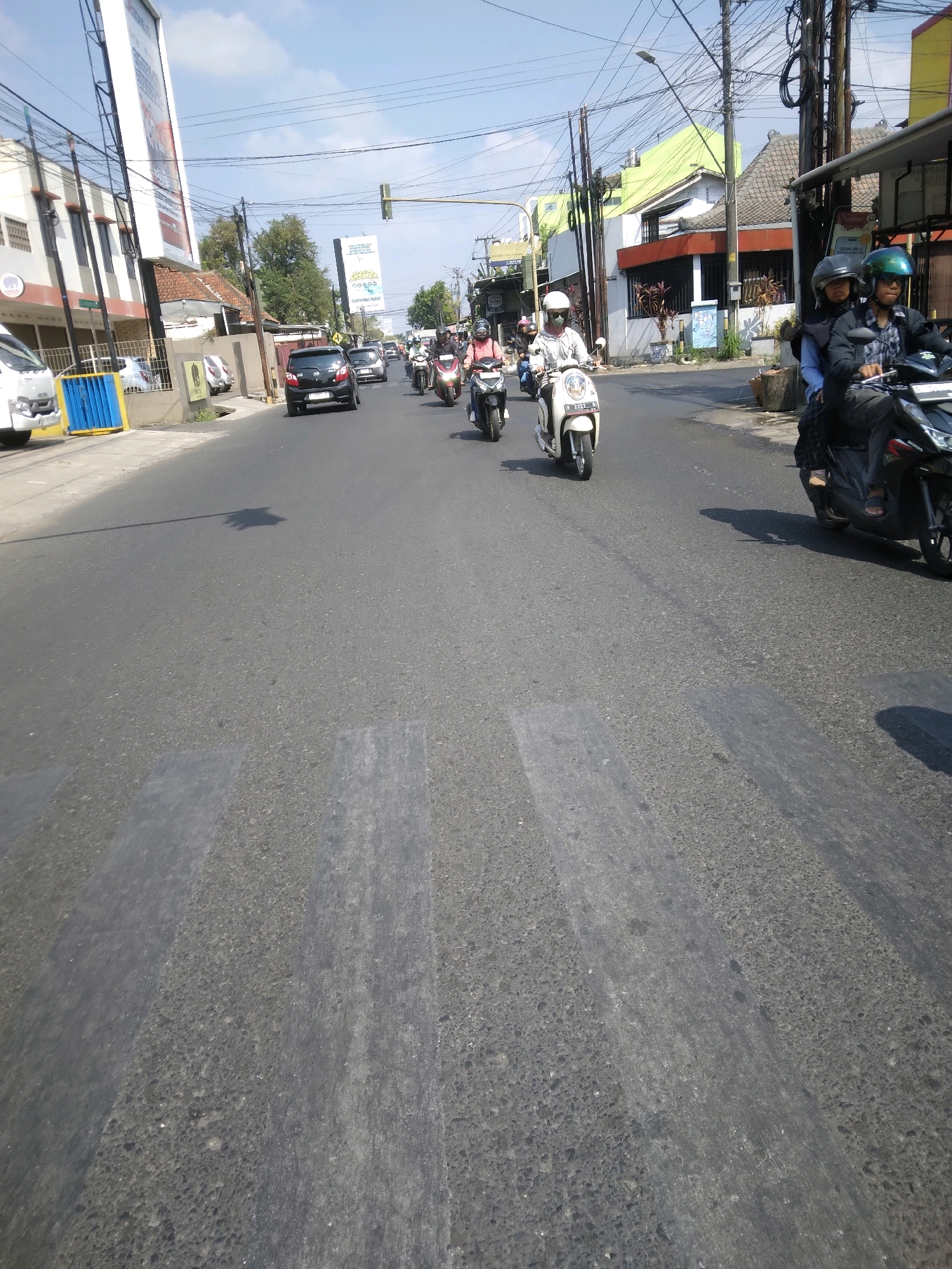 This is a street-level, eye-level shot of a busy street in Purwokerto, Indonesia. The scene is dominated by a paved road with a pedestrian crossing marked by light gray stripes. Several motorcycles, each carrying one or two people, are traveling along the road. The riders are wearing helmets, and some passengers also have helmets on. On the right side of the road, a dark-colored motorcycle with two riders is prominent. In the mid-ground, a white scooter with a single rider is moving forward. Further up the road, other motorcycles and a black car are visible. The buildings lining the street are varied in style, some featuring balconies and colorful paint, suggesting a commercial and residential area. Overhead, a tangle of electrical wires crisscrosses the sky, and traffic signals are visible in the distance. The weather appears to be clear and sunny, with a bright blue sky.
