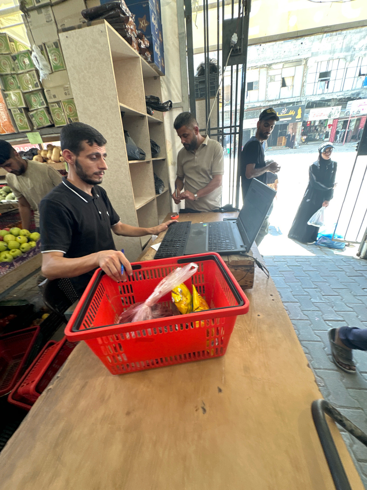 A man in a black polo shirt with white trim is standing behind a wooden counter, interacting with a laptop. In front of him is a red plastic shopping basket containing plastic-wrapped items. To his left, another man in a beige short-sleeved shirt is partially visible, looking down. In the background, shelves are stocked with boxes and bags. Beyond the open doorway, a street scene is visible with buildings and a woman in dark clothing holding a plastic bag. The scene appears to be a market stall in Gaza City, Palestinian Territories.