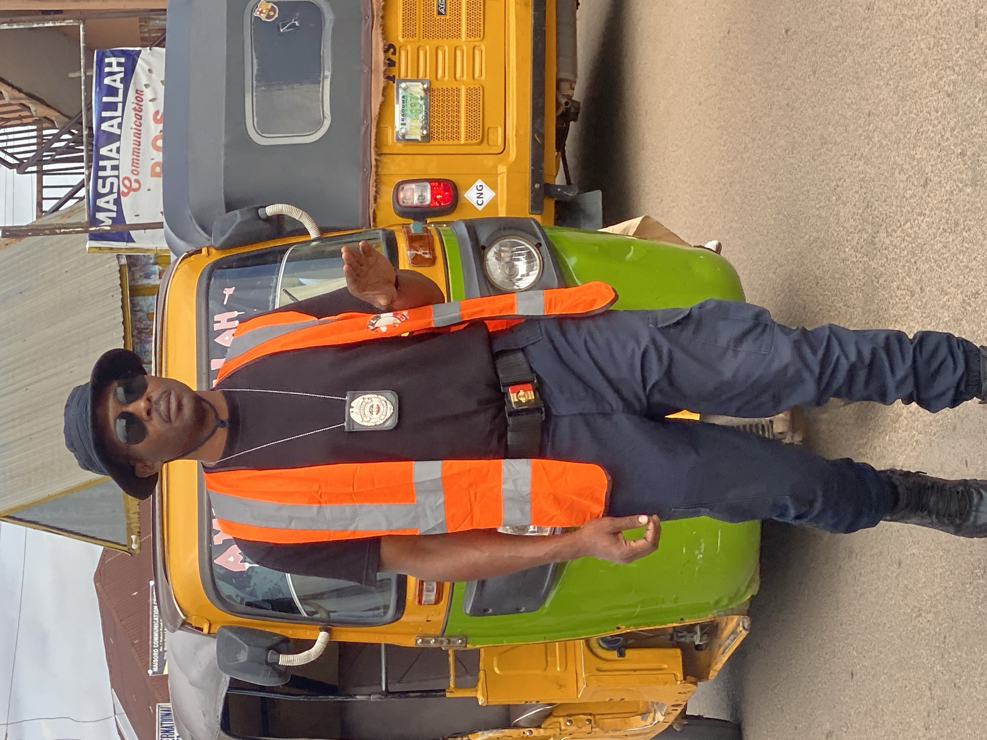 In Rigachikun, Nigeria, a Black man wearing a black bucket hat, dark sunglasses, a black t-shirt, an orange safety vest with reflective stripes, dark pants, and black boots stands on a street next to two auto-rickshaws. He has a badge hanging from a lanyard around his neck. One auto-rickshaw is yellow and green, and the other is gray. The man is gesturing with his right hand, palm facing outward. In the background, there are buildings and signs, including one that reads "MASHA ALLAH Communication POS." The street is paved with asphalt.
