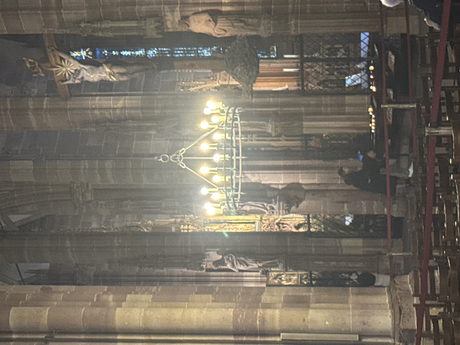 The image displays the interior of the Cathédrale Notre-Dame de Strasbourg, France, characterized by grand Gothic architecture. Tall, segmented stone columns and arches frame the scene under diffused lighting. Centrally, a large circular metal chandelier with approximately sixteen lit electric candle-shaped bulbs hangs. To the upper right, a monumental crucifix features a sculpted figure of Christ with a sunburst halo, set against a narrow, vertically oriented stained-glass window displaying blue and white hues. Stone statues of robed figures are integrated into the architectural elements; one stands prominently on the right, and another is partially visible on the far left. Gilded decorative screens are discernible in recessed arches. In the midground, two individuals are present near a low, dark stand holding multiple lit votive candles. One person, dressed in a dark jacket and light trousers, faces the candle stand, while another, wearing a dark coat, stands slightly behind. The foreground contains rows of empty wooden pews with red upholstered seats, separated by red ropes strung between stanchions. The environment is dimly lit, with the chandelier and votive candles providing warm light, creating atmospheric highlights and shadows.