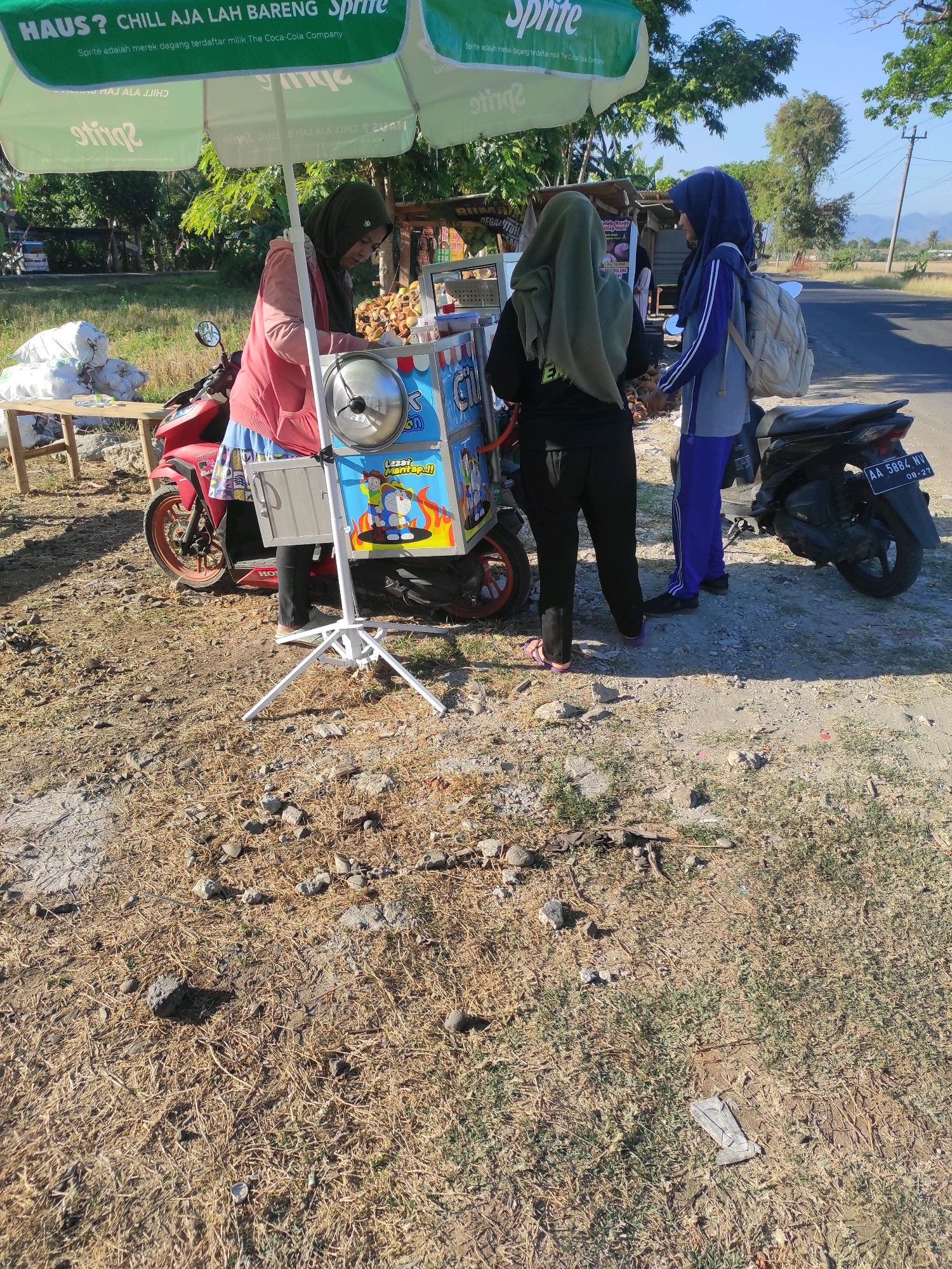 Three women are standing next to a food cart under a green umbrella advertising Sprite. The woman on the left, wearing a pink jacket and headscarf, appears to be serving food. The woman in the middle, wearing a grey headscarf and black clothing, is facing away from the camera. The woman on the right, wearing a blue headscarf and blue and grey athletic wear, is facing the woman in the middle and has a backpack. A red motorcycle is parked to the left of the food cart. The scene appears to be outdoors on a dirt road next to a paved road, with trees and vegetation in the background. The food cart is decorated with cartoon characters. The license plate on the motorcycle reads "AA 5884 NV 08-27". The location is identified as Purwodadi, Indonesia.