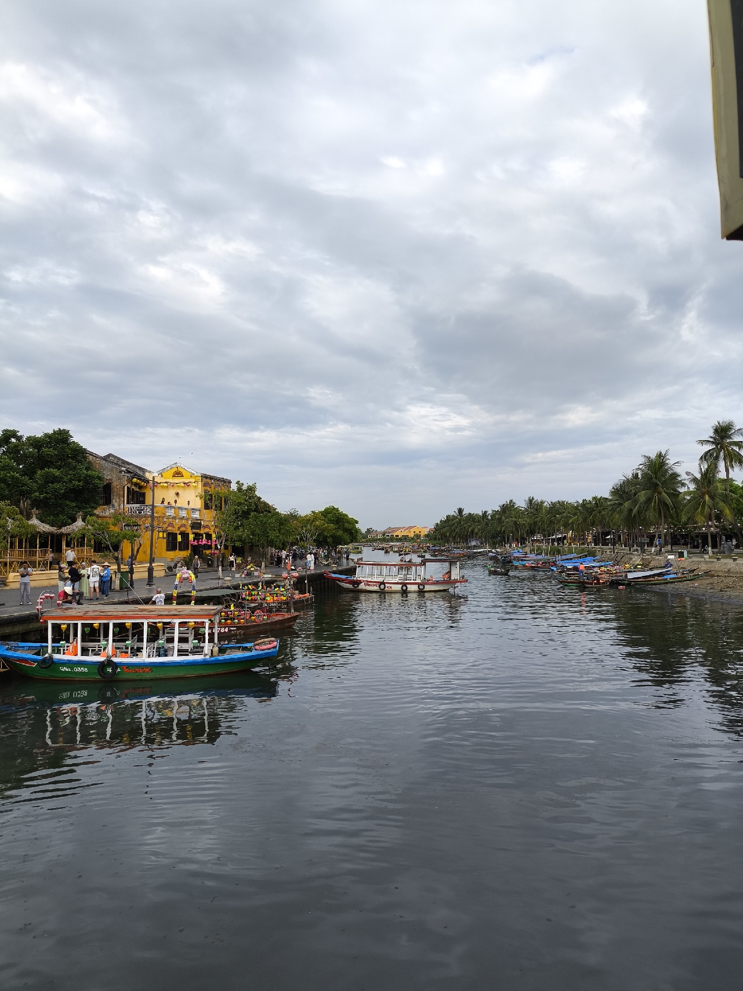 This is a medium-shot, vertical, eye-level, full-color photograph of a river scene in Hoi An, Vietnam, on an overcast day. The foreground is dominated by the dark, rippling water of the river.
In the middle ground, several boats are moored along the river. The most prominent boat in the left foreground is a tourist boat, painted green and blue with an orange roof and white railings. It has several rows of seats and life vests visible. The number "QNA 0358" is visible on its side. Behind it, other boats of varying sizes and colors, some appearing to be tourist boats with canopies, are also docked.
The background features a picturesque street scene. On the left bank, there are traditional yellow and ochre-colored buildings with wooden balconies and tiled roofs, characteristic of Hoi An's ancient town. Lush green trees and vegetation line the riverbank, with palm trees visible further down the bank. People are walking along the riverside promenade, some dressed in casual clothing, others in what appear to be traditional conical hats. Small stalls and awnings are set up along the walkway, likely for vendors.
On the right bank, more boats are moored, and a line of palm trees forms a dense backdrop. The sky is filled with a heavy blanket of grey clouds, suggesting a likely chance of rain. The overall atmosphere is tranquil, depicting a typical day along the Thu Bon River in Hoi An.