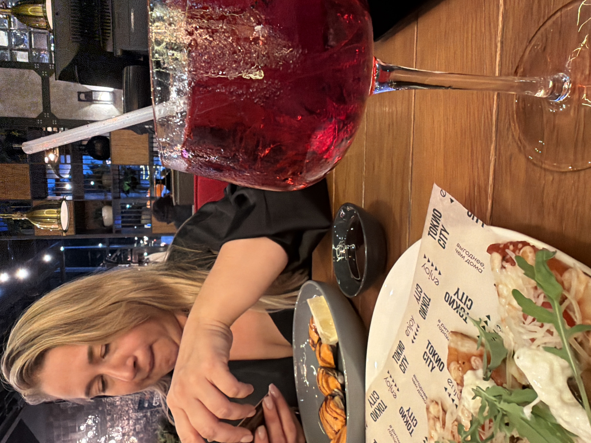 A woman with blonde hair is seated at a wooden table in a restaurant. She is wearing a black top and is focused on her food, which appears to be sushi. To her right, a large glass of red beverage with a straw is prominently displayed. On the table in front of her, there is a plate with sushi and a small bowl of sauce. Further to the right, a pizza is partially visible on a white paper liner. The restaurant's ambiance is suggested by the background, which includes shelving, lighting fixtures, and what appears to be city lights visible through windows. The overall scene captures a moment of dining in a modern establishment.