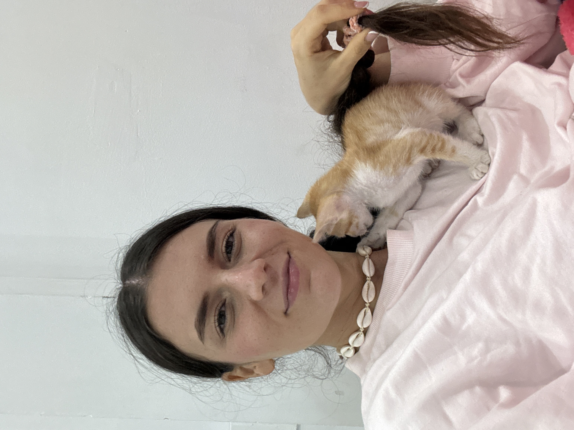 A young woman is depicted in a close-up, portrait-style shot, gently holding a small kitten. The image is oriented sideways, with the woman's head and upper torso angled towards the top.
The woman has dark hair, which she holds up with her left hand, securing it with a light pink scrunchie. Her dark eyebrows frame a smiling face, and she looks directly towards the viewer with a warm, affectionate expression. Her complexion is clear with visible freckles. She is wearing a light pink, long-sleeved top or sweater, and a distinctive necklace made of white cowrie shells adorns her neck.
Nestled comfortably on her shoulder or upper chest is a small, ginger and white kitten. Its back is facing the camera, with its hind legs and tail visible, suggesting it is resting or sleeping.
The background is a plain, light-colored (white or off-white) wall, indicating an indoor or simple, clean setting. The lighting is bright and even, suggesting it is daytime. There is no visible text in the image. The scene conveys a sense of calm and tender interaction between the woman and the kitten, presumably in Mũi Né, Vietnam.