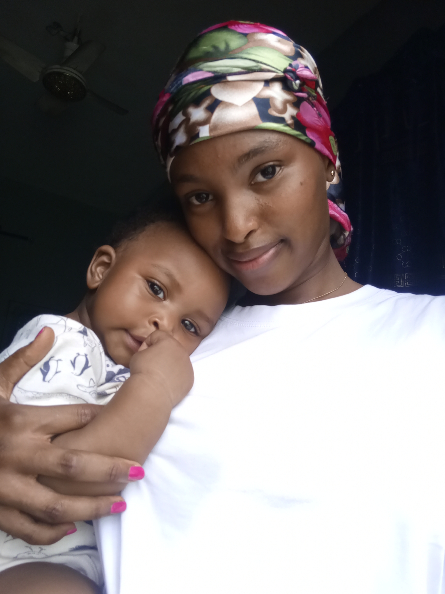 The image presents a close-up, intimate portrait of a woman holding a baby in an indoor setting. Both individuals have dark skin and are positioned centrally, with their faces illuminated by a light source coming from the front, while the background remains dim.
The woman, likely the mother, is wearing a plain white crew-neck t-shirt and a colorful headscarf featuring patterns of green, pink, brown, and black. She has a delicate gold chain around her neck and small gold stud earrings. Her fingernails are painted a vibrant fuchsia pink. She looks directly at the camera with a gentle, loving gaze and a faint, soft smile, conveying warmth and affection.
Cradled in her left arm and resting against her chest is the baby, who appears calm and content. The baby is wearing a white long-sleeved onesie with small dark patterns, possibly animals or abstract shapes. The baby's head is slightly tilted, and its eyes are wide and curious as it also looks towards the camera. One of the baby's hands is raised to its mouth.
The background is mostly obscured by shadows. In the upper left corner, a cream-colored ceiling fan is dimly visible. To the right, a dark curtain or wall adds to the muted backdrop. The overall mood of the photograph is one of tenderness, love, and peaceful connection between the woman and the child. There is no visible text in the image.