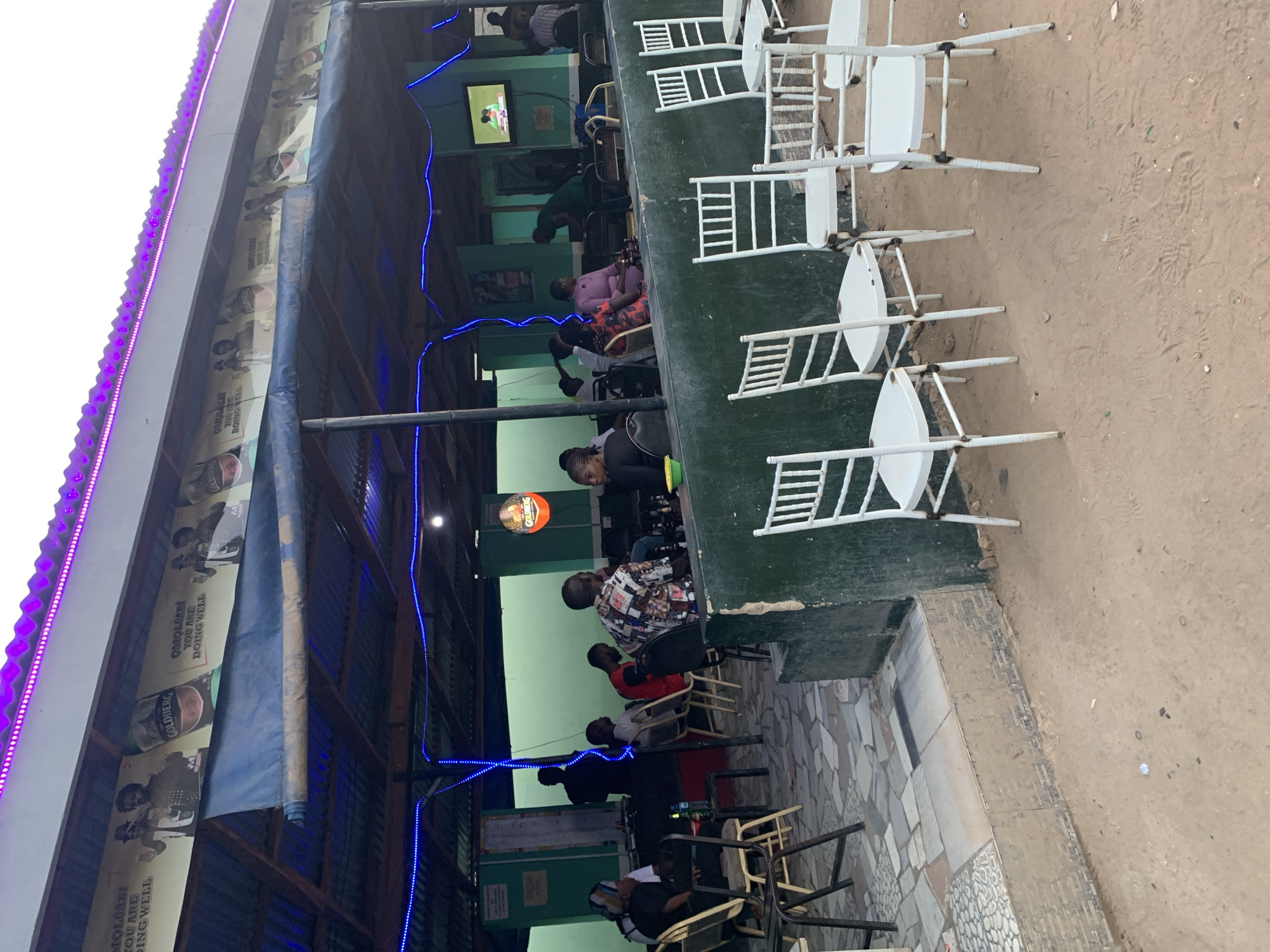 The image shows an outdoor seating area of a bar or restaurant in New Stadium, Nigeria. The establishment has a partial roof adorned with advertisements for "Goldberg" beer and blue LED lighting. The seating consists of white plastic chairs and some metal chairs, arranged along a green elevated platform. Several people are seated, some watching a television mounted on the wall. The ground adjacent to the seating area is dirt.