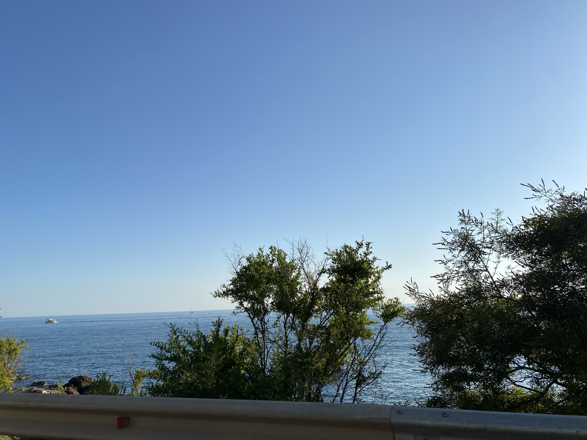 The image depicts a serene coastal scene in Sutomore, Montenegro. The foreground features a weathered metal guardrail, consistent with roadside barriers, with a red and white marker visible on its surface. Beyond the guardrail, lush green vegetation, including trees and shrubs, partially obscures the view of the azure Adriatic Sea. The sea stretches to the horizon under a clear, bright blue sky, with a few small boats visible on its surface, suggesting a calm day. The overall impression is one of natural beauty and tranquility, likely captured from a vantage point overlooking the coastline.