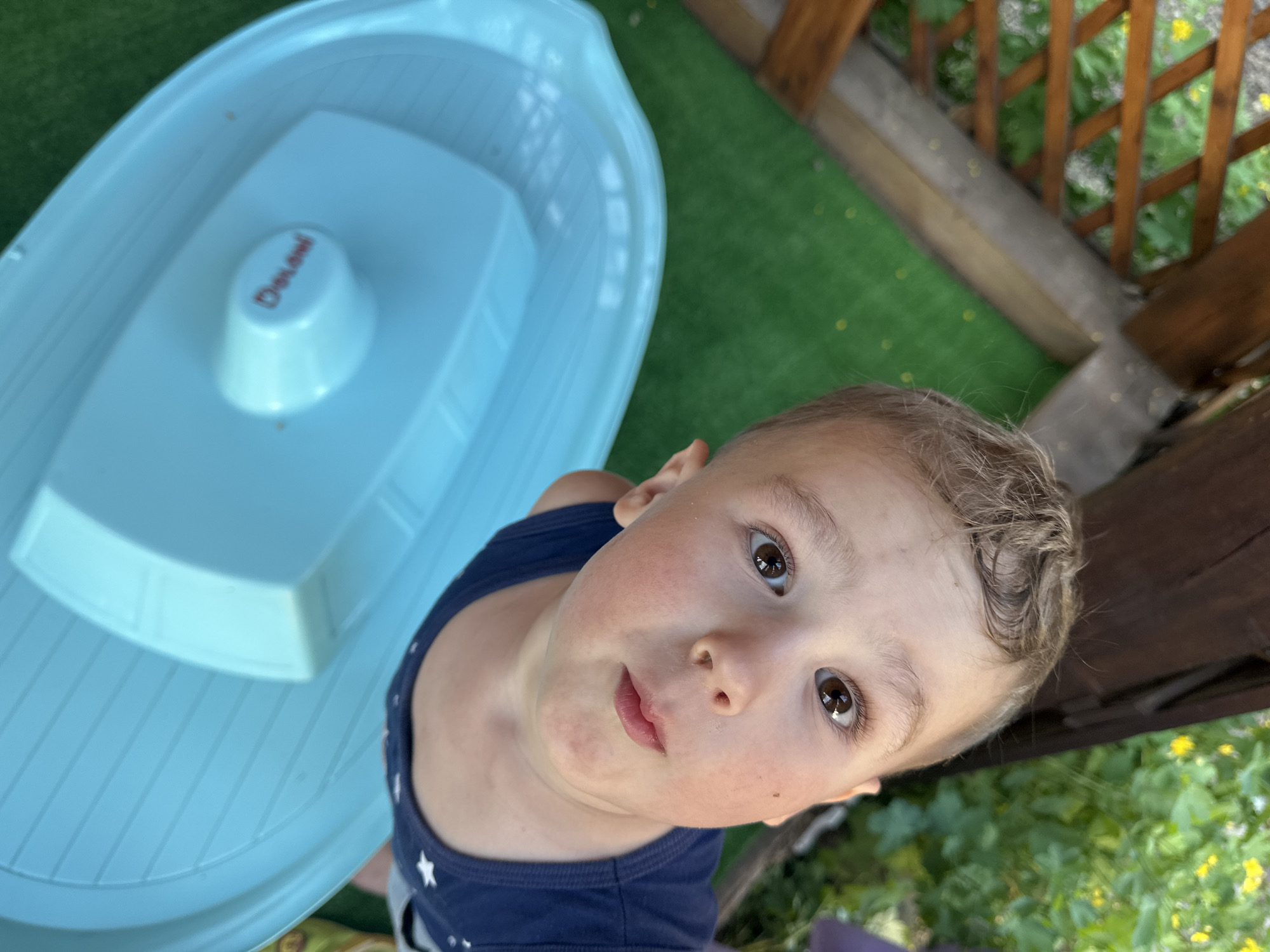 A high-angle shot shows a young boy looking upwards, positioned below the camera's view. The boy has fair skin, brown eyes, and light brown hair. He wears a navy blue tank top with white stars. Adjacent to him is a light blue, plastic boat-shaped tub. The tub is on a green surface. Wooden beams and green foliage form the background. There is no indication of the location being Nizhny Novgorod, Russia.