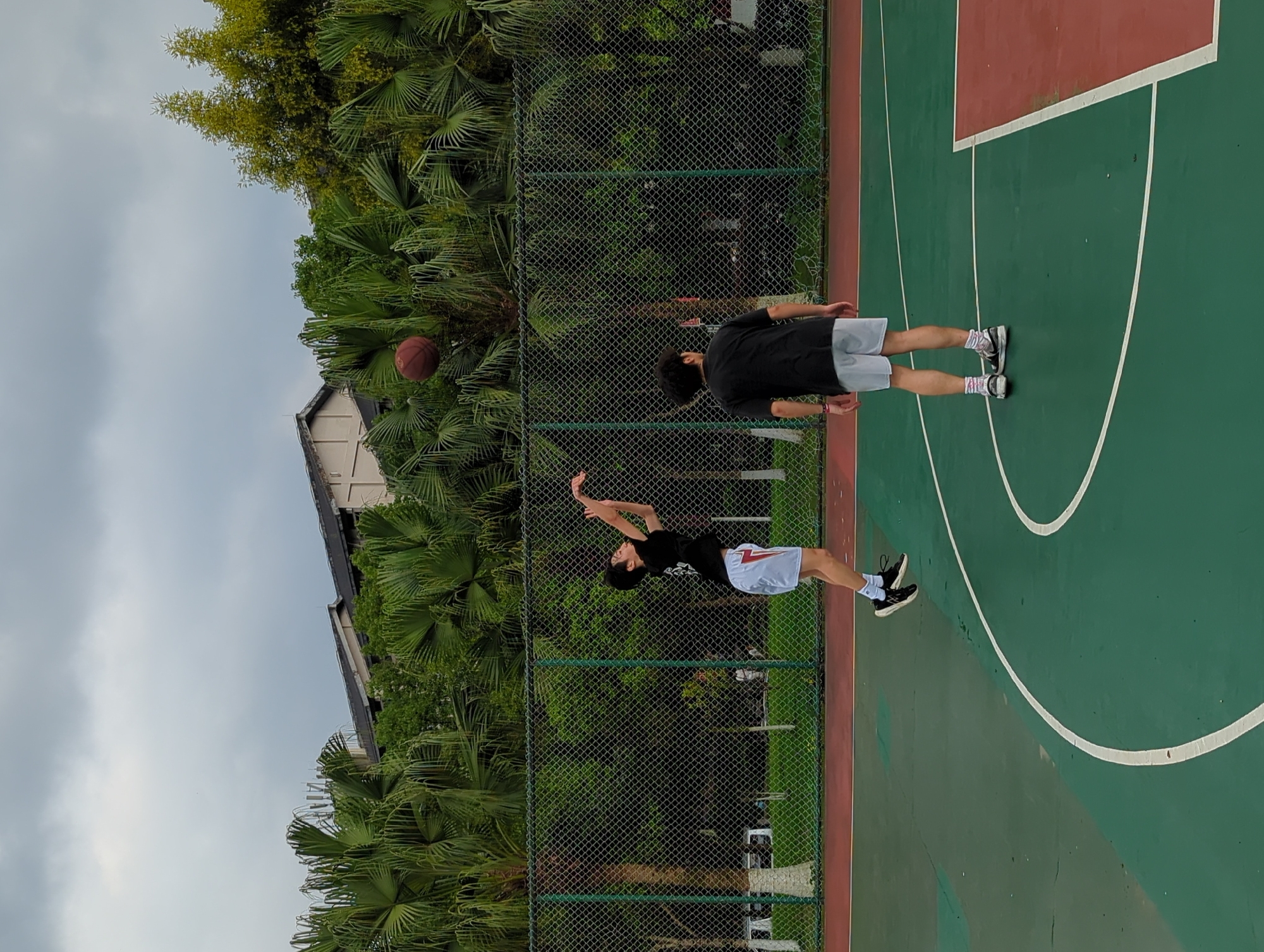 A basketball game is in progress on a green and red court. Two males are present; one in a black shirt and white shorts is about to shoot a basketball, while another male, also in a black shirt but with grey shorts, is observing. The basketball is in mid-air, slightly left of the shooter. A wire fence is visible behind the players. Beyond the fence, there's a dense collection of trees and partial view of a building under a cloudy sky. The location is suggested to be in the city of Jiangjin, China.