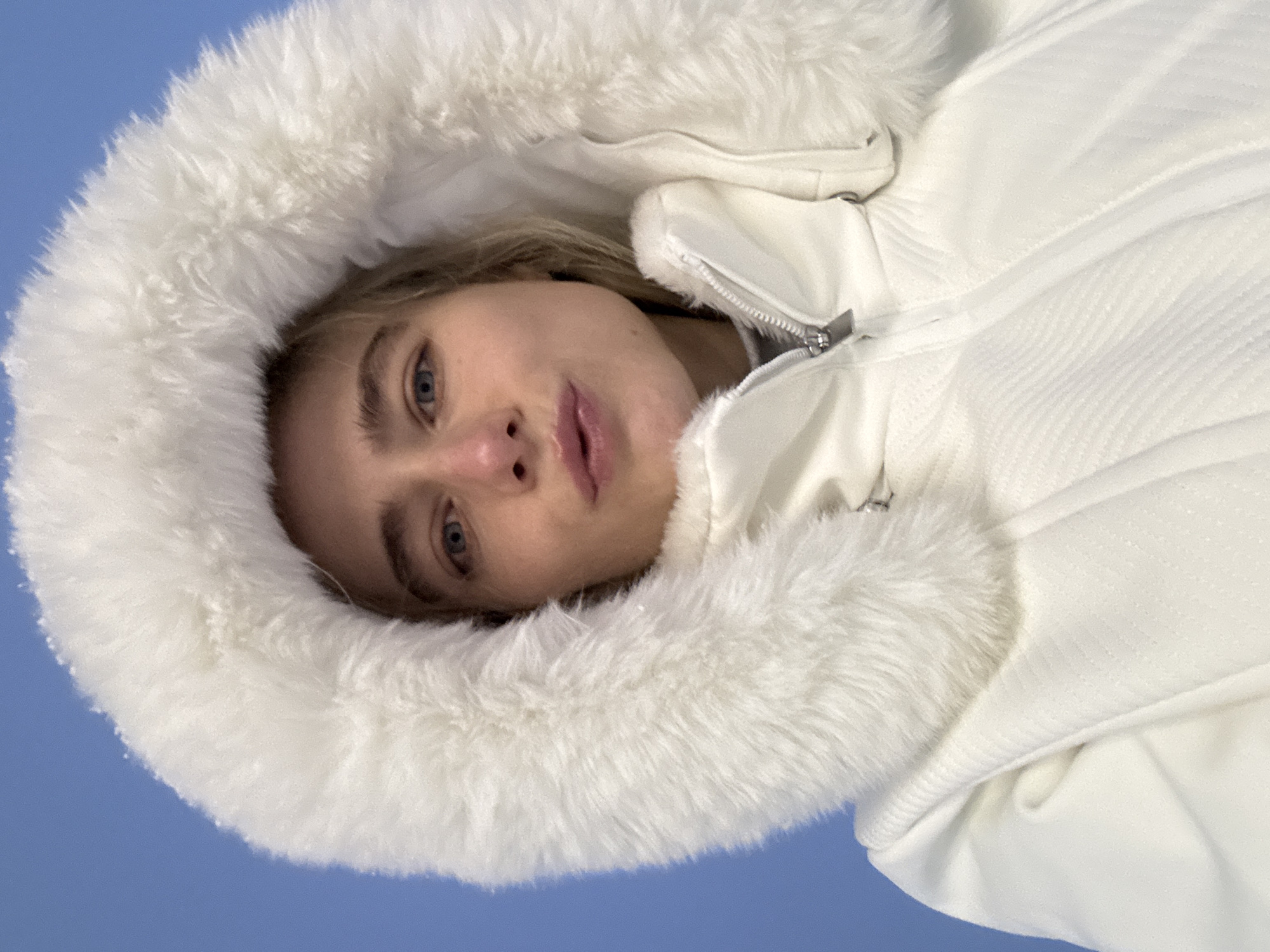 A person with light skin and blue eyes, wearing a white puffy jacket with a fur-lined hood, is looking directly at the camera. The background is a solid blue, suggesting an outdoor setting, perhaps under a clear sky. The jacket is zipped up and appears to be made of a quilted material. No information about the city or location is available.