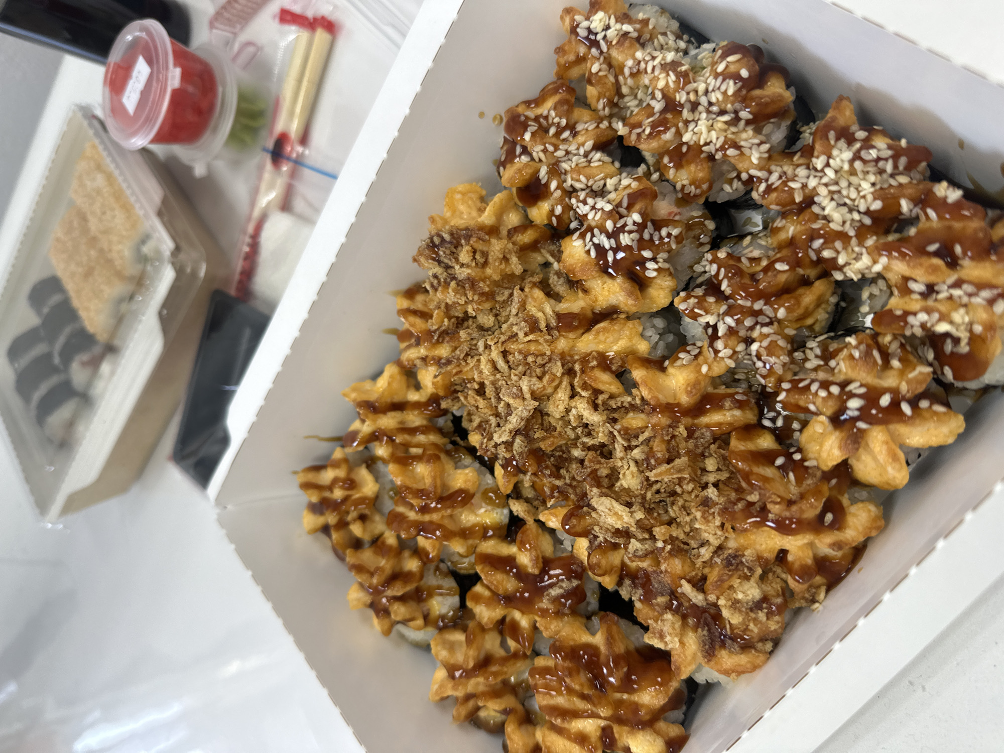 A high-angle, close-up shot shows a white takeout box filled with sushi. The sushi is decorated with fried tempura, brown sauce, and sesame seeds. A plastic container holding a red sauce and chopsticks is partially visible to the left of the box. Another container, presumably containing more sushi, is also visible on the left. The background is a blurry white surface. It is unclear where this image was taken or whether it is from Votkinsk, Russia.