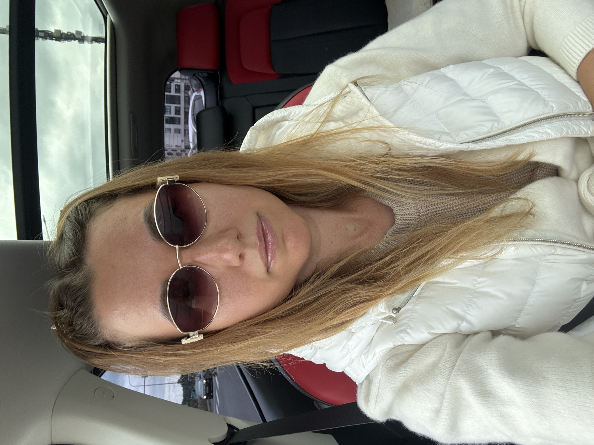 A medium shot shows a fair-skinned woman with blonde hair pulled into a ponytail and wearing gold-rimmed sunglasses. She is seated in the passenger seat of a car, her body angled slightly to the left. She is wearing a white quilted vest over a cream-colored sweater. The car has a panoramic sunroof, and through it, a gray, cloudy sky is visible, along with some buildings and a cell tower. The interior of the car features red and black seats. The woman is looking directly at the camera.