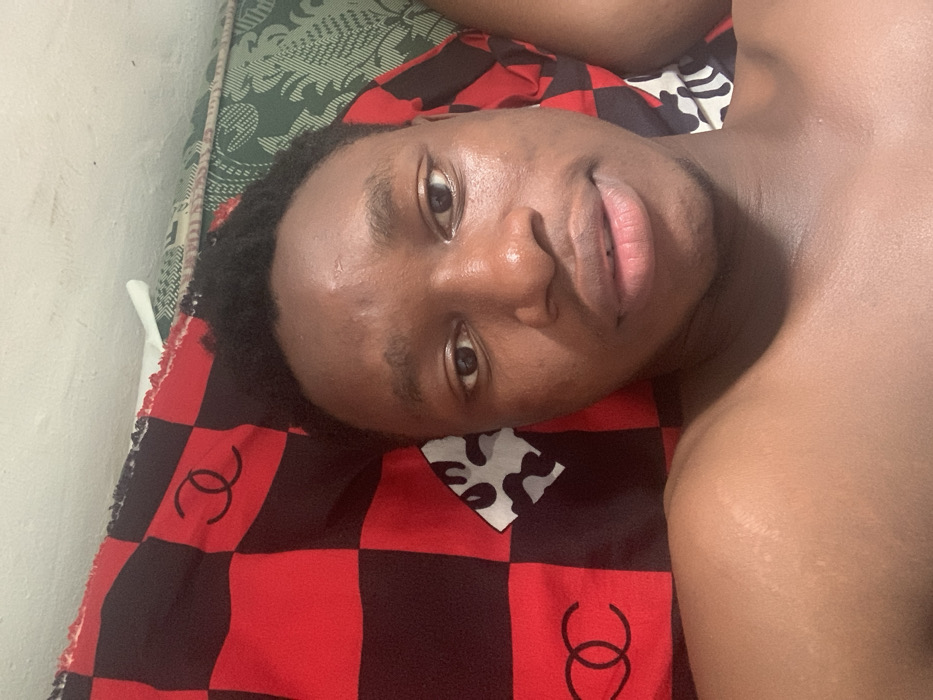 An indoor scene in Eziobodo, Nigeria, depicts a young adult male taking a selfie while lying down. The individual, of dark complexion with short dark hair, is positioned with his head resting on bedding. He looks directly at the camera with a neutral expression, his lips slightly parted. His bare upper chest and shoulders are visible. To the left side of the frame, a red and black checkered fabric with a black double-C logo resembling the Chanel brand is present. To the right, a green fabric with an intricate pattern is visible. A section of a light-colored wall is observable at the top of the frame. Another fabric featuring abstract white/grey and black shapes is visible near the subject's right shoulder.