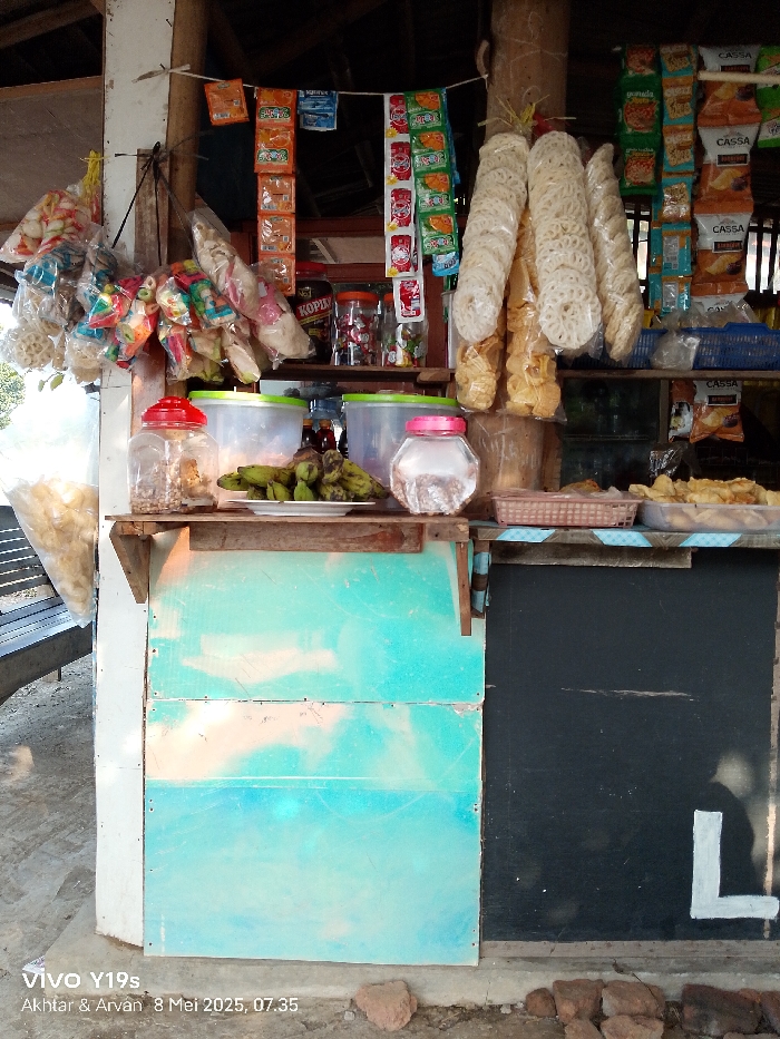 The image shows a small, open-air shop in Kecamatan Anjatan, Indonesia. The shop is located at coordinates -6.42 latitude, 107.978 longitude. The shop is made of wood and has a blue and black painted surface behind the counter. The counter is covered with a variety of packaged goods, including snacks, candy, and drinks. There are also several glass jars containing food items. The scene is likely taken during the daytime, as there is no artificial light present. There are no people in the image. There are several baskets and containers full of crackers and other snacks. The shop is closed as there are no people in the image and the shop is not actively serving customers. The shop is located in an urban environment, as evidenced by the brick flooring and the neighboring buildings in the background.
