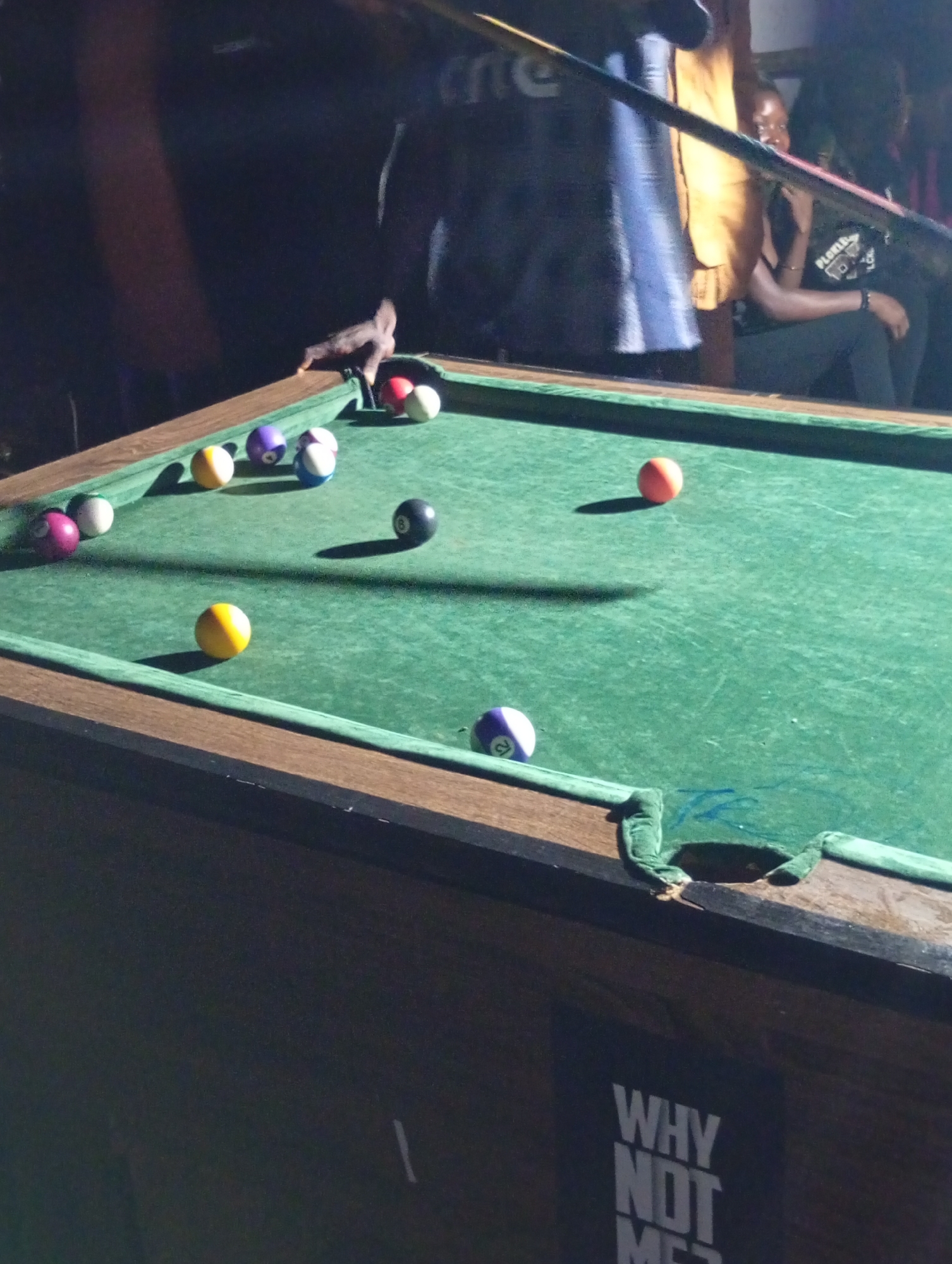 A group of people are gathered around a green felt billiards table at night, in what appears to be an indoor establishment in Durumi, Nigeria. Several billiard balls are scattered across the table, including a red ball, a yellow ball, a purple ball, a ball with a black "8" on it, and a ball with "12" on it. A pool cue is positioned over the table, and a hand is visible, seemingly in the process of taking a shot. The lighting is dim, casting shadows on the table and illuminating parts of the individuals present. Some people are seated and others are standing. The overall atmosphere suggests a recreational gathering.