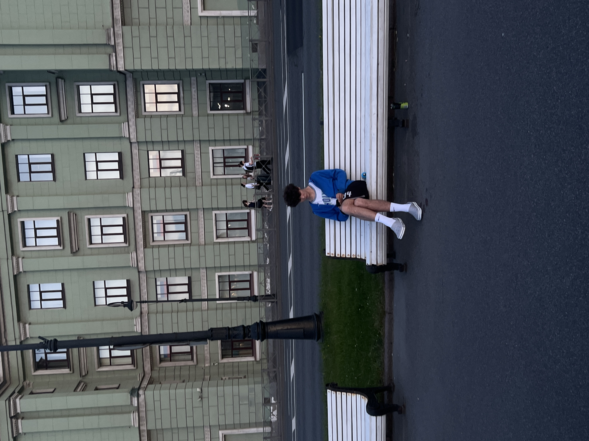 A young person with dark curly hair sits alone on a white slatted bench, holding a mobile phone in their hands. They are wearing a blue hooded jacket over a white t-shirt and dark shorts, with white socks and grey slip-on shoes. The bench is situated on a paved area, with a strip of green grass and a black lamppost behind it. In the background, a light green building with many windows stands, and a road with a sidewalk and railing is visible. Several people are walking on the sidewalk in front of the building. The scene appears to be in a city, possibly during the late afternoon or early evening.