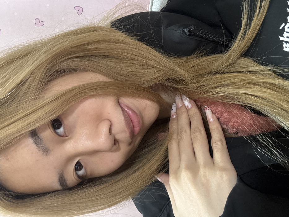 A medium close-up, front-facing portrait displays a young adult East Asian female in an indoor setting. She has light brown hair, dark eyes, and a subtle smile with glossed lips. She wears a black puffer jacket, revealing a section of a pink, fluffy garment underneath. Her right hand, featuring long, almond-shaped fingernails with elaborate nail art (white abstract designs, red polka dots, and a small red bow), is positioned on her upper chest area, resting on her clothing. The background consists of a light pink wall adorned with scattered pink heart outlines and small white dots. The specific location within Shapingba District, China, cannot be determined from the visual information provided.