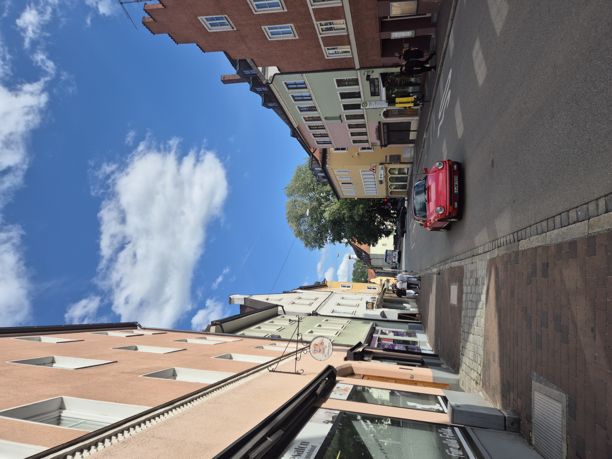 The image depicts a street scene in Landshut, Germany, specifically the Altstadt. The viewpoint is from a pedestrian's perspective on the left side of the street, looking down the street as it curves slightly to the right. The street is lined with colorful, historic buildings, characteristic of the Altstadt. The buildings are multi-story, with distinct architectural styles, featuring pitched roofs with dormer windows, arched doorways, and a variety of window shapes and colors. The facades are painted in pastel shades of peach, green, yellow, and pink, contrasted by a prominent reddish-brown building on the right side of the image.
The street is paved with asphalt, and there is a brick sidewalk on the left side, bordered by cobblestones. Various shops and businesses are visible on the ground floor of the buildings, indicated by signage and storefronts. One storefront on the left displays text that includes "für alle Felle," suggesting a pet-related business. Another sign, hanging from a bracket, features a circular graphic of a dog. Further down the street, a sign reads "Jimmy Barber Shop."
In the center of the street, a red Porsche car is driving towards the viewer. The car has its headlights on and a German license plate that reads "LA HH 911H". Several pedestrians are visible on the sidewalk. On the left sidewalk, three individuals are walking away from the camera. On the right sidewalk, two figures are walking towards the camera, and further down, a group of three people are walking away from the camera. A white bus is parked or driving on the right side of the street in the distance.
The sky is bright blue with scattered white clouds, indicating fair weather. A large tree with dense foliage is visible between the buildings on the left side of the street, further down. The overall impression is that of a charming, historic European city street on a sunny day.