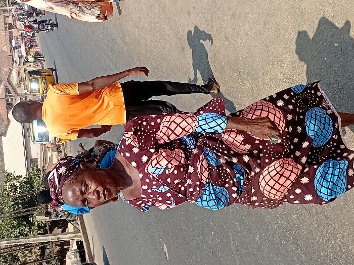 An elderly woman in a patterned dress and headwrap stands in the street, holding a small object. Behind her walks a younger man in an orange shirt and dark pants. A yellow vehicle is visible in the background, along with buildings and other people. The scene is lit by bright daylight, casting shadows on the pavement.