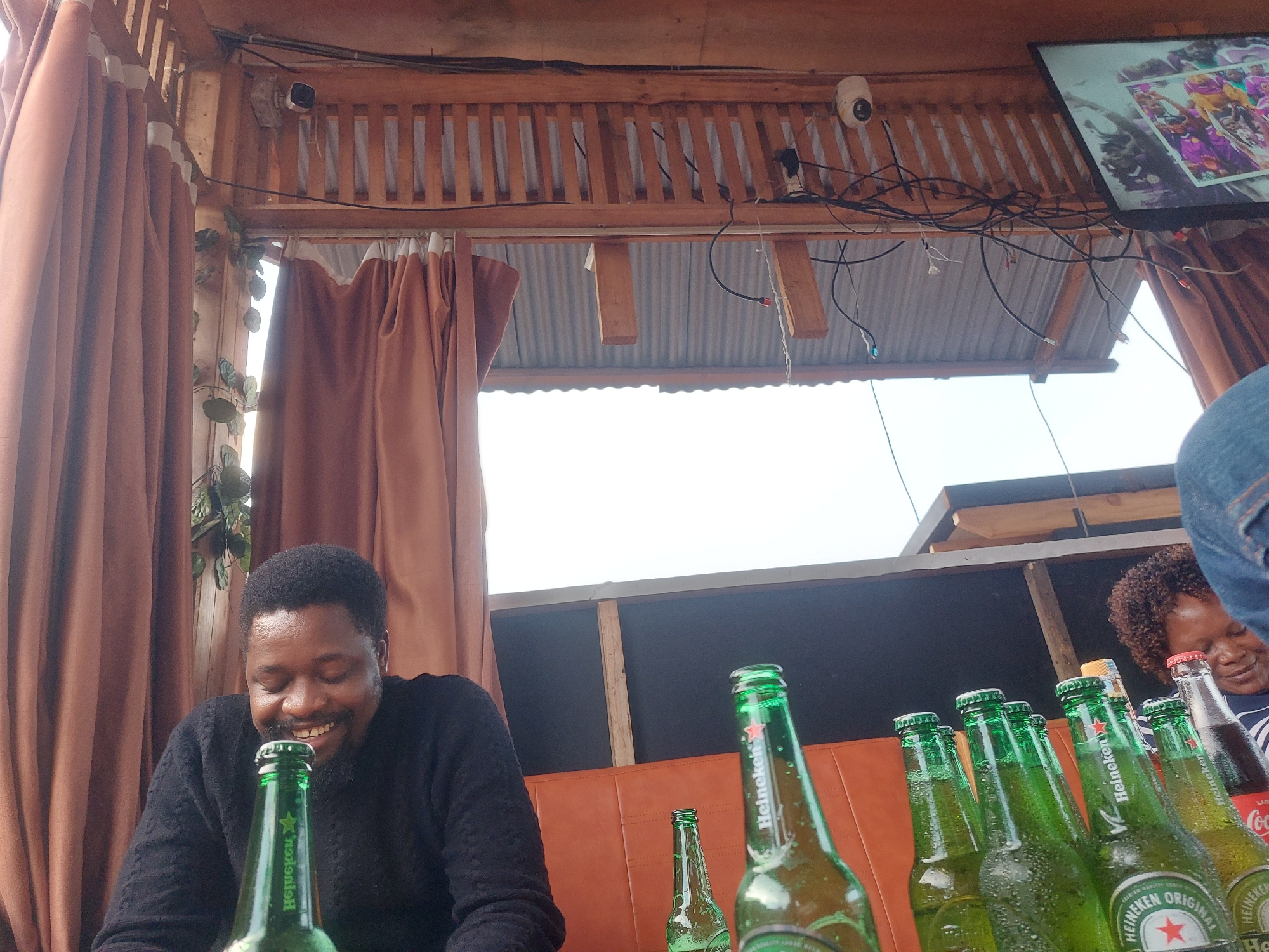 The image captures a casual outdoor setting, likely a bar or restaurant in Dar es-Salaam, Tanzania. An African man wearing a black sweater is smiling behind a row of green Heineken beer bottles on a brown leather seat. A woman, partially visible, is looking at the beer bottles. A TV is mounted overhead in the background. The area is covered with a wooden structure with brown curtains.