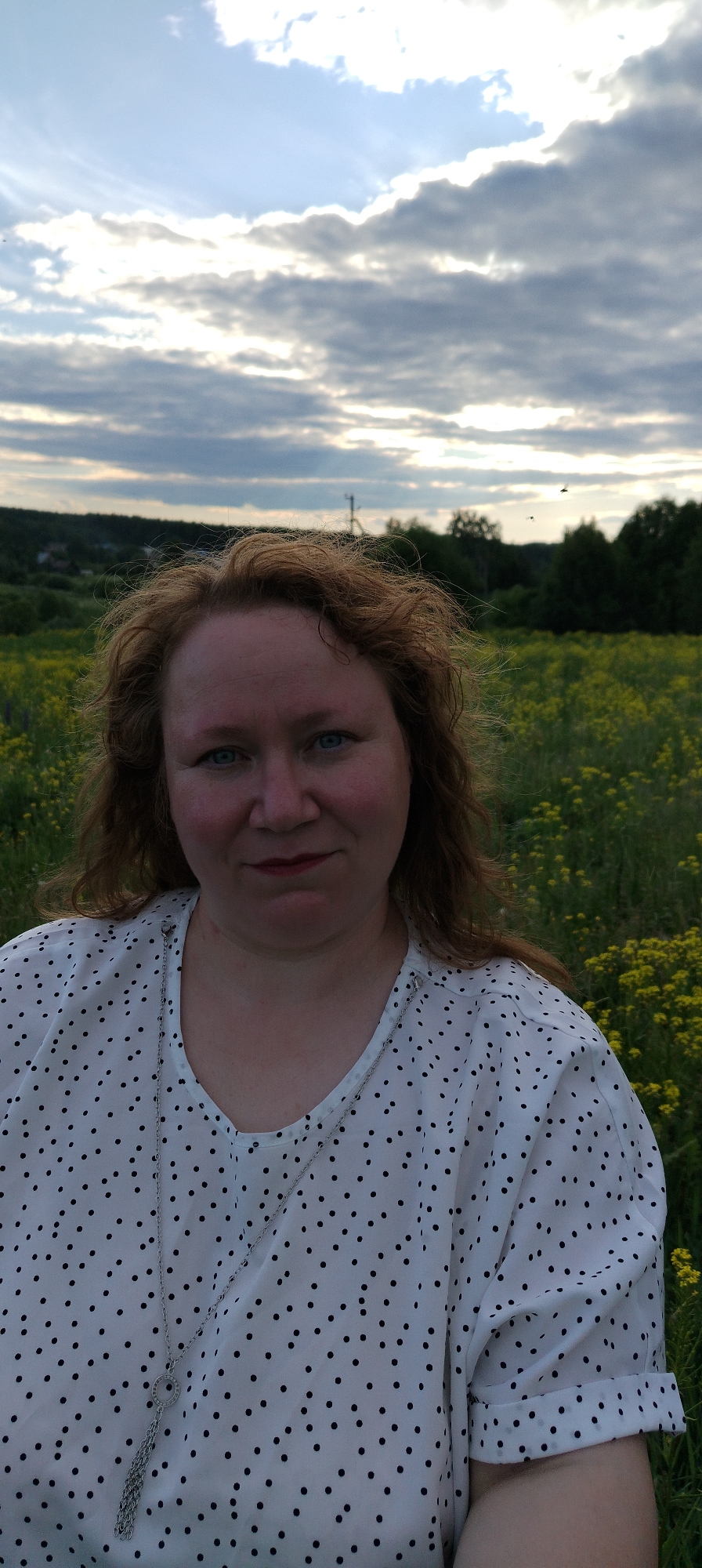 The image captures a woman outdoors in Зипуново, Russia. She is positioned centrally, looking directly at the viewer with blue eyes and a slight, neutral smile. Her reddish-brown, wavy hair appears somewhat disheveled or windblown. She is wearing a white blouse adorned with numerous small black polka dots and a long silver-colored necklace featuring a circular pendant with decorative elements and multiple dangling chains. The immediate background shows a field densely populated with small yellow flowers and green vegetation. Further in the background, a line of dark green trees forms a dense wooded area. To the left, distant structures resembling houses are visible, and a utility pole extends above the treeline. The sky is largely overcast with a mix of gray and white clouds, revealing some brighter patches and areas of lighter blue. Several small, dark specks, possibly birds, are visible against the sky in the upper right portion of the image. The lighting is soft and diffused, consistent with an outdoor scene under cloudy conditions.