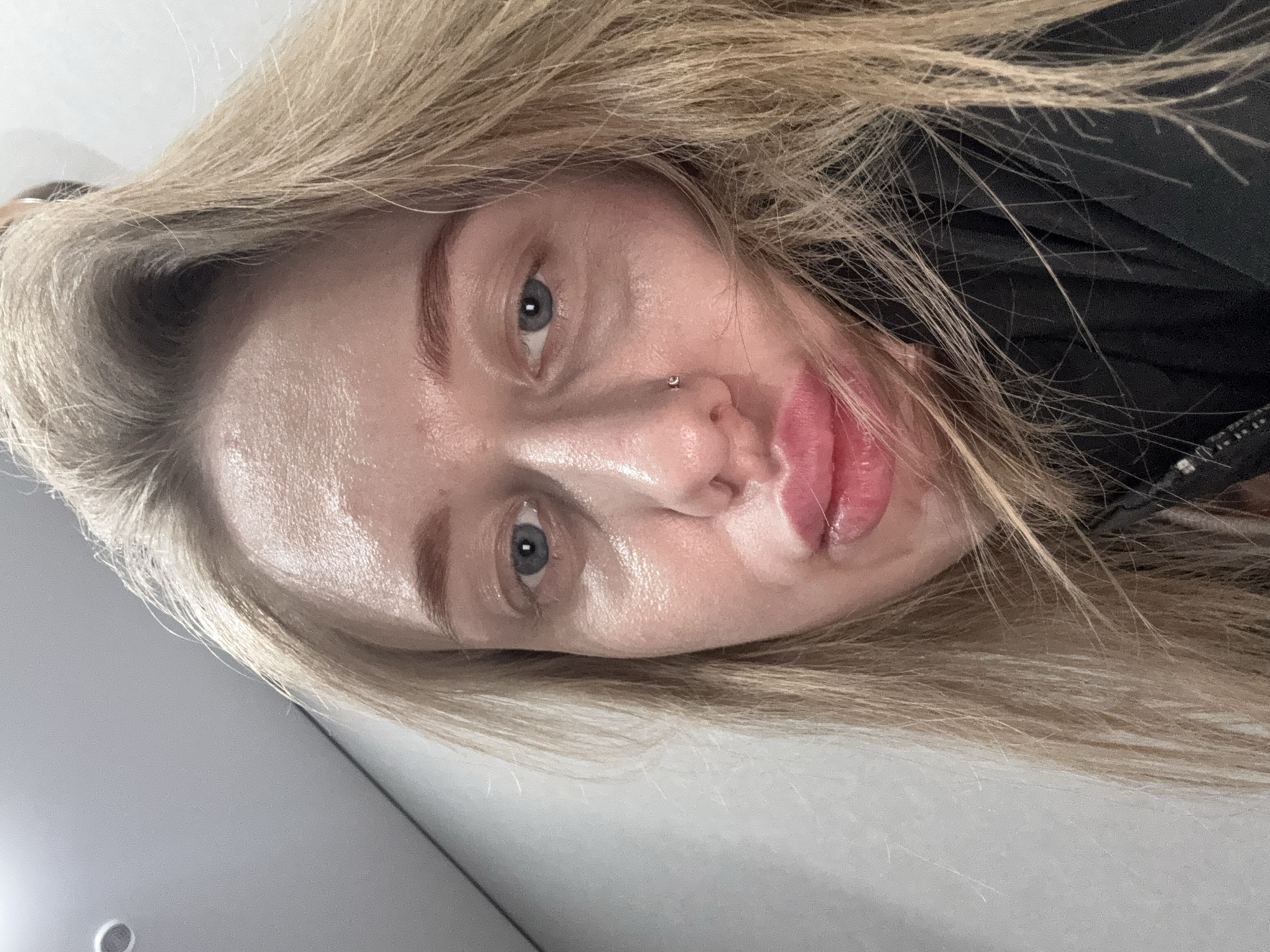 A close-up, tilted, high-angle shot depicts a woman with fair skin, blonde hair, and light blue eyes. She has a piercing in her left nostril. Her lips appear full and slightly parted. Some strands of her blonde hair are draped across her face. She is wearing a dark-colored garment. The background is predominantly out of focus and appears to include a light grey surface. The image contains no indication of the city of Rostov-on-Don, Russia.