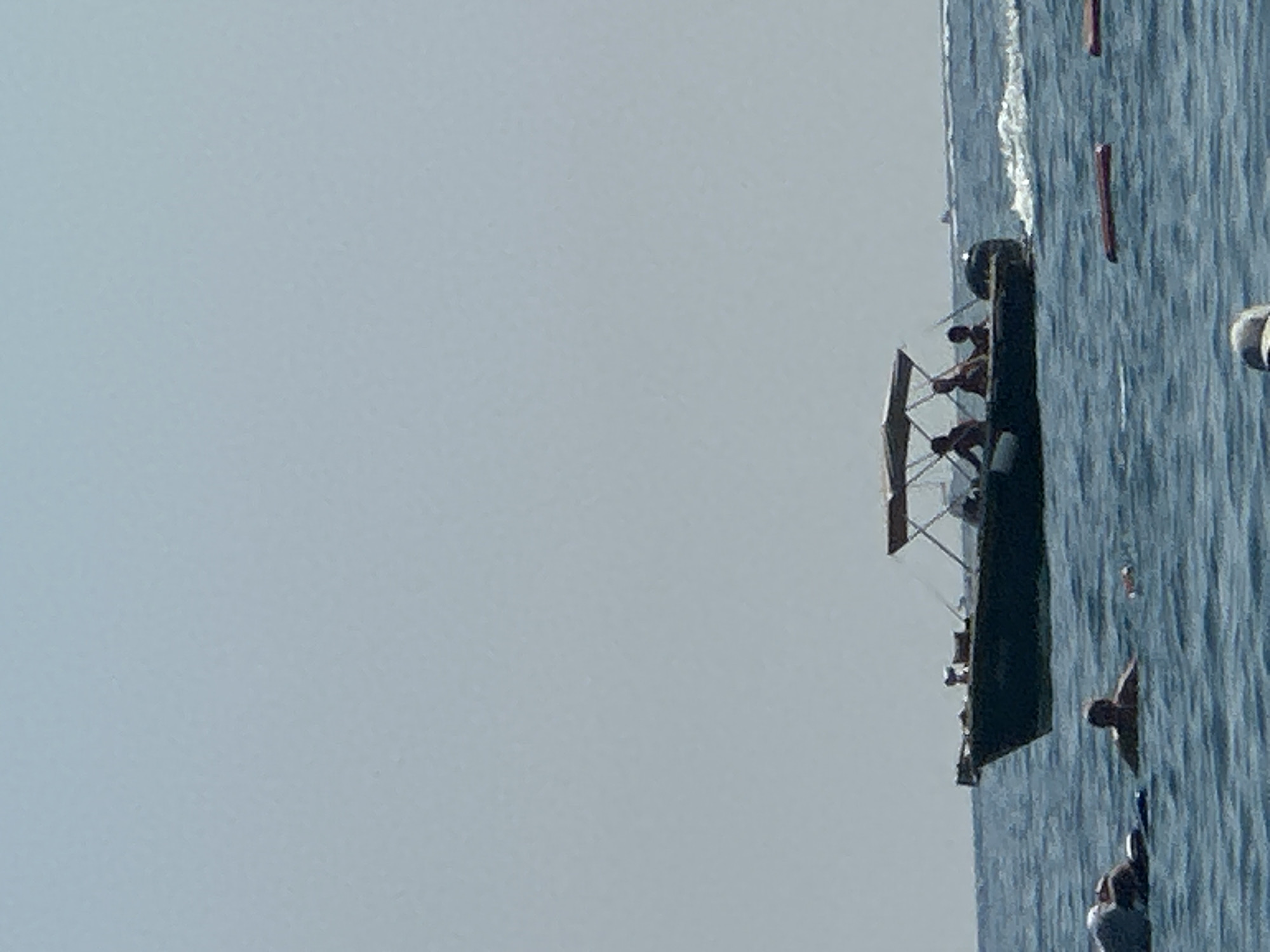 A wide-angle, eye-level shot captures a serene coastal scene in Boreti, Montenegro, under a clear, pale blue sky. The water, a deep, undulating blue, fills the majority of the frame. In the mid-ground, a dark-hulled motorboat with a canopy sails across the water, leaving a white wake in its path. Four people are visible in the boat, their figures silhouetted against the bright sky. The boat appears to be moving from left to right, away from the viewer.
In the foreground, several individuals are enjoying the water. To the left, two people are partially submerged, one lying on their back and the other with their head visible above the surface. Further to the right, another person is swimming, with their shoulders and head visible. A few red floating objects are scattered in the water between the boat and the swimmers. The overall atmosphere is one of leisure and recreation on a sunny day.