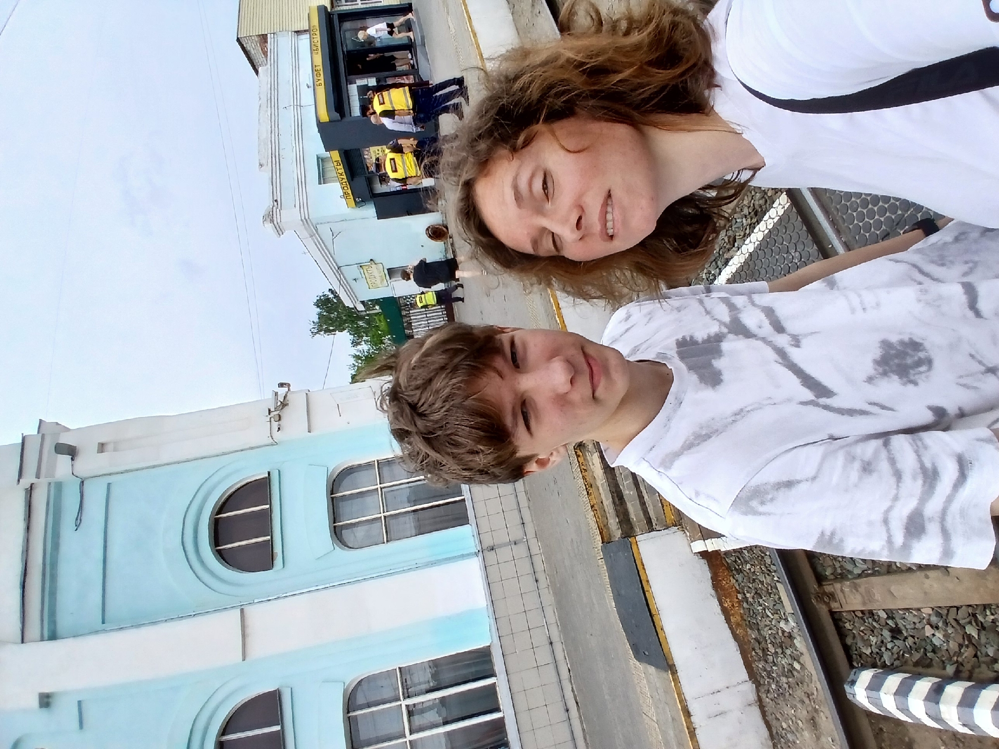 This is a selfie taken at the Rtishchevo railway station in Rtishchevo, Russia. Two individuals are in the foreground, a young male with light brown hair wearing a tie-dye t-shirt, and a woman with curly brown hair wearing a white t-shirt. They are standing near train tracks. In the background, there is a light blue and white building with arched windows, characteristic of a station. Further in the background, there is a small building with yellow signage that reads "ПРОДУКТЫ" (Products) and "БУФЕТ «БИСТРО»" (Buffet "Bistro"). Several people are visible near this building, including some wearing bright yellow vests, likely railway personnel. The sky is overcast.