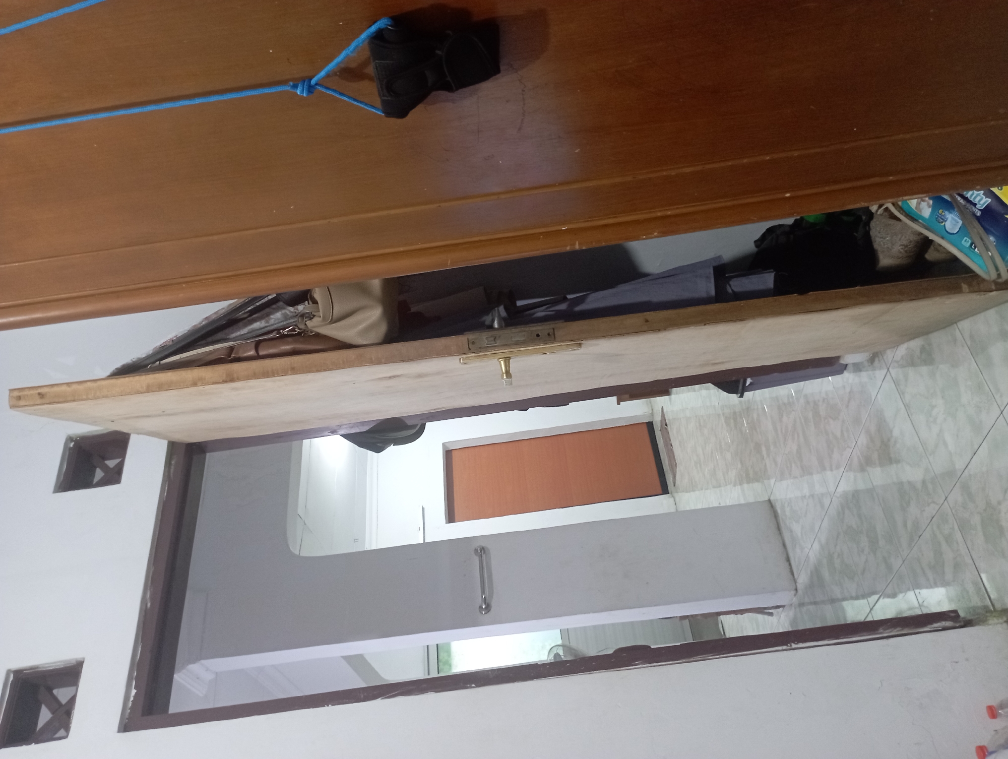 The image shows an interior hallway with two open doors and a wooden door on the right side of the frame. The hallway has tiled flooring and white walls. The first door on the left leads to another room with a second door visible further down the hall, which is orange. A chrome towel bar is mounted on the white wall in the first room. The open door in the center of the frame is made of unfinished wood and reveals a closet area with clothing and bags. On the wooden door to the right, a black object is hanging by a blue rope. The scene appears to be inside a residential or commercial building in Purwakarta, Indonesia, given the architectural style and typical interior design elements found in the region.