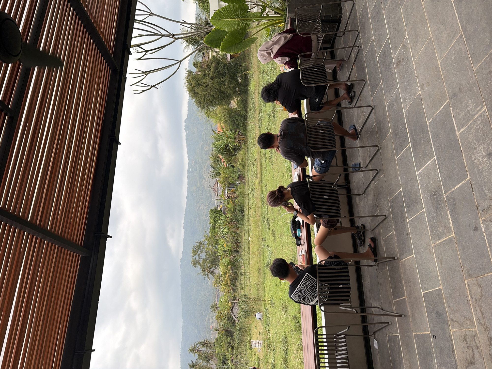 Five individuals are seated at a table with a view of a rural landscape in Buleleng, Indonesia. The individuals are positioned from left to right: a person facing away from the camera, a person on their phone, a person on their phone and possibly eating, a person eating, and a person wearing a headscarf and eating. The table is a long, dark wooden surface. The seating area is covered by a slatted wooden roof with visible metal supports. In the background, a green field with scattered trees and a distant treeline of hills under a cloudy sky is visible. The foreground consists of a stone-tiled patio. A ceiling fan is mounted on the underside of the roof structure.