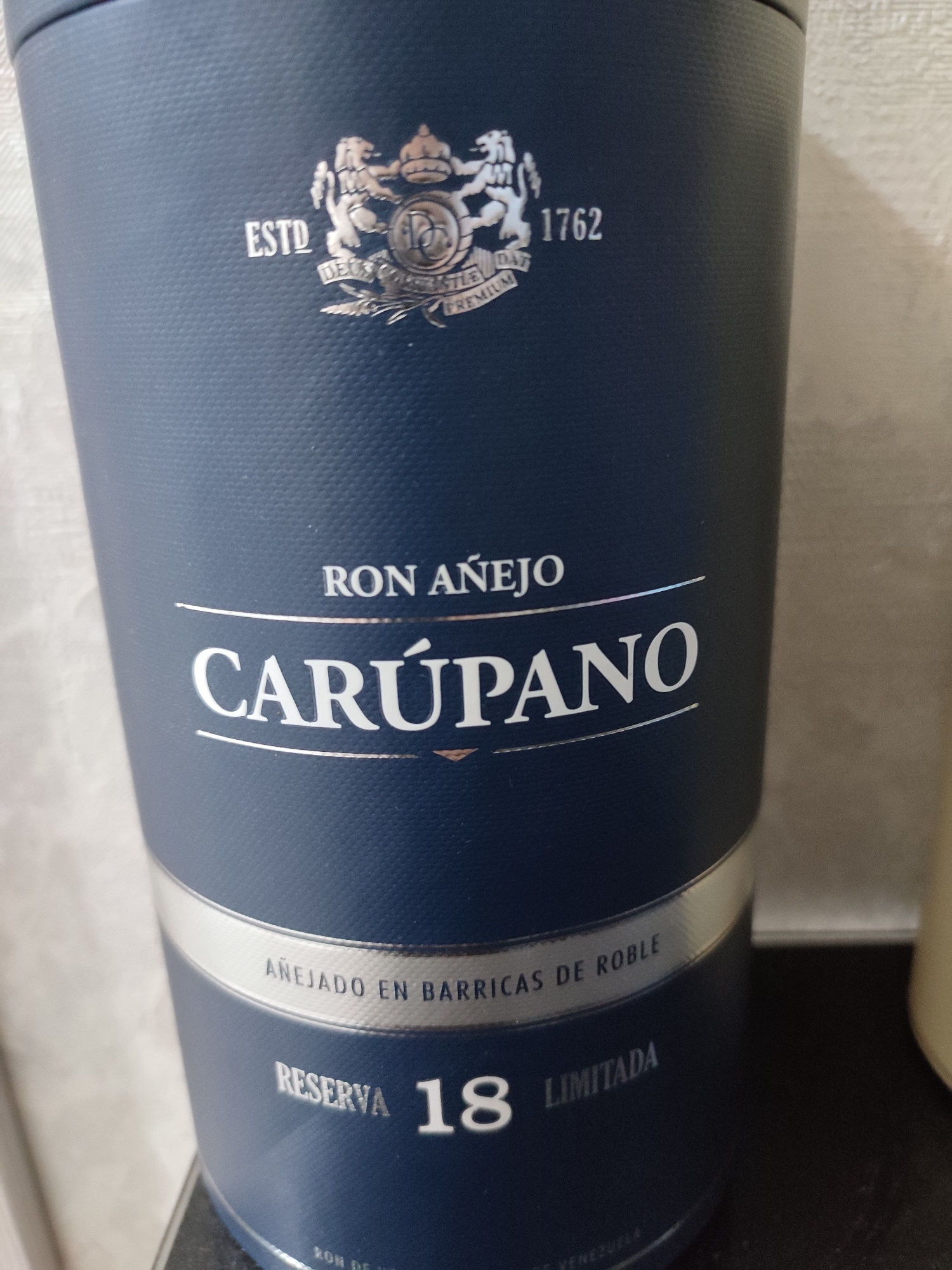 An image displays the side of a dark blue cylindrical bottle. The bottle has embossed text and a crest. The crest features a lion and a crown. To the left of the crest, it reads "ESTD". To the right of the crest, it reads "1762". Below the crest, text is partially visible, including "DEUX" and "PREMIUM". Below this, "RON AÑEJO" is written. Prominently displayed below that is "CARÚPANO". Further down, a silver band circles the bottle with the text "AÑEJADO EN BARRICAS DE ROBLE". Below this band, "RESERVA 18 LIMITADA" is visible. At the very bottom, faint text reads "RON DE VENEZUELA". The bottle is set against a plain, light-colored background.