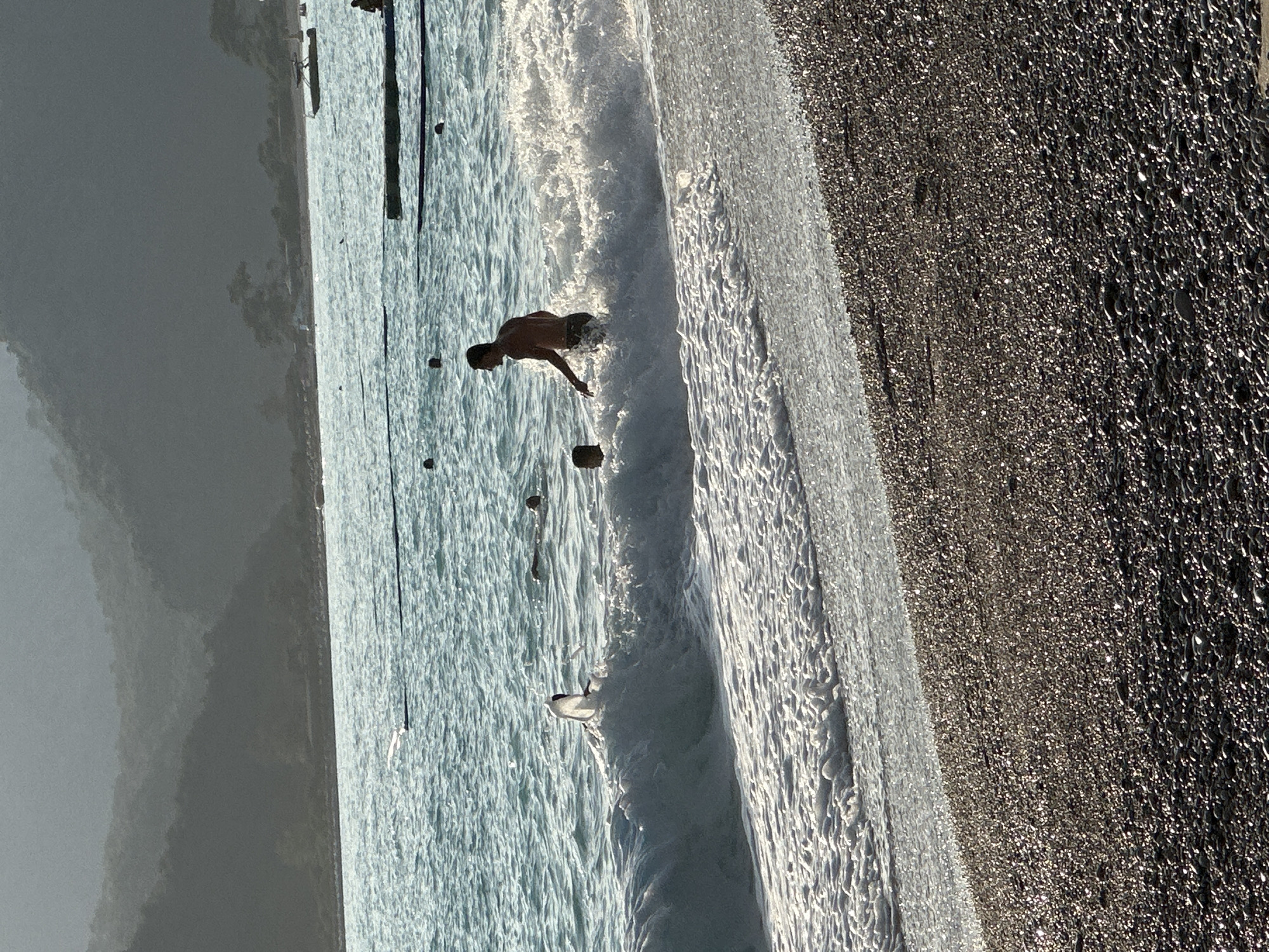 A man stands in ocean surf beside a beach in Fethiye, Turkey. The beach is on the right and is made of small, dark stones. Waves break onto the shore. The man, shirtless and wearing dark shorts, is partially submerged in the water. In the distance, there is a mountainous terrain under a grey sky. Wooden pylons protrude from the ocean. A bird floats in the water near the shoreline.