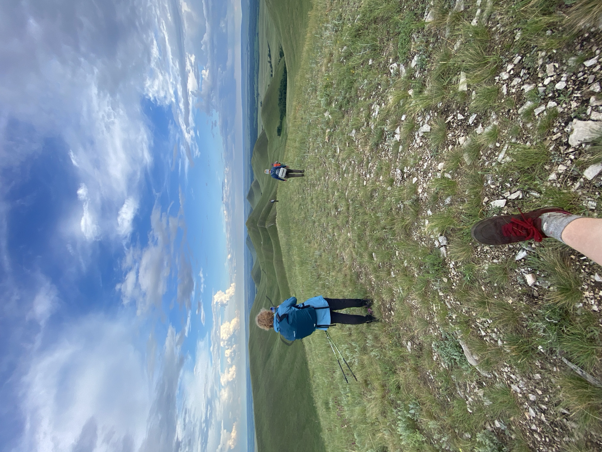 The image depicts a wide, rolling landscape with undulating green hills under a cloudy blue sky. In the foreground, a person's leg wearing a brown hiking shoe and grey socks is visible. Further into the scene, three individuals are hiking along a grassy ridge. The person closest to the viewer, wearing a blue jacket and backpack, is looking at their phone. Another person, in the middle distance, is walking away from the camera, and a third hiker is visible in the far distance. The terrain is covered in grass and scattered rocks. The location is identified as the city of Verkhneozernoe, Russia.