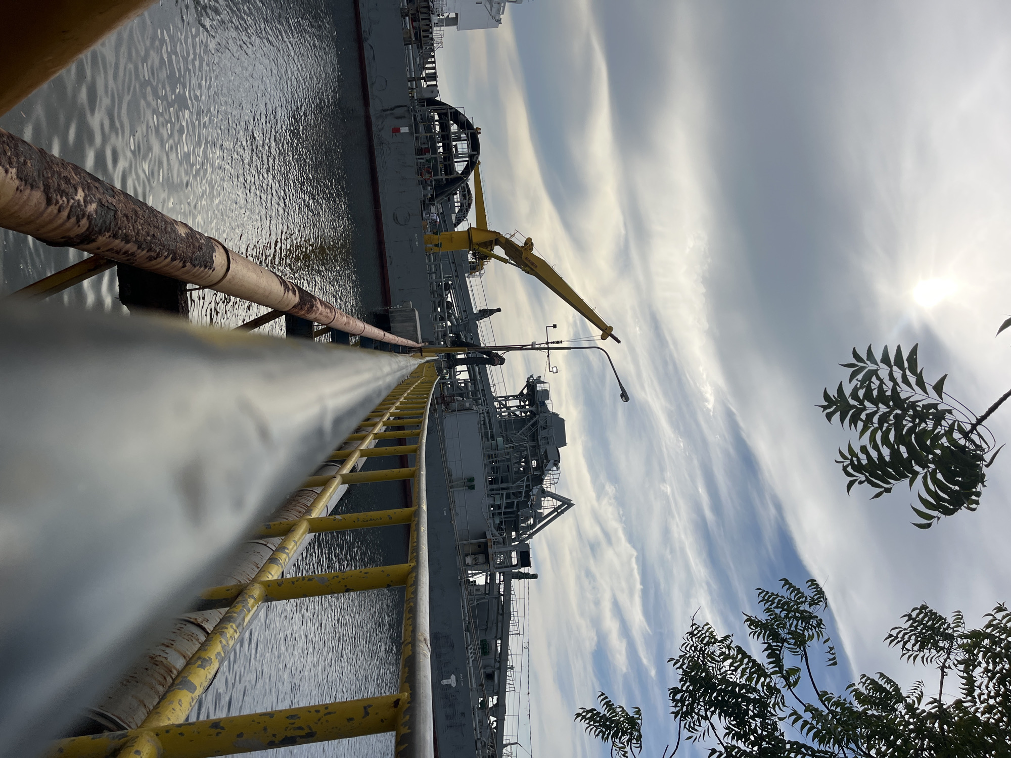 The image depicts a scene at a port facility in Tambonbaroh, Indonesia. A large gray ship is docked alongside a concrete pier. Yellow metal pipes and a ladder run parallel to the water's edge in the foreground. A yellow crane is visible on the ship's deck. The water is calm and gray. The sky is overcast with wispy clouds and the sun is visible. Trees with dark green foliage are visible in the lower right corner of the frame. The overall impression is of an industrial or commercial maritime operation.