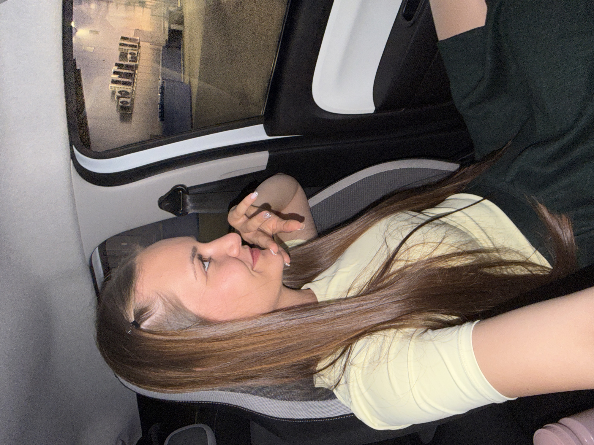 A young woman with long, straight, brown hair is seen reclining across the front passenger seat of a car. She is wearing a light yellow long-sleeved top. Her head is turned towards the camera, and she is looking gently to the side with a soft, thoughtful expression. Her right hand, adorned with light-colored nail polish, is raised near her face. Her hair is spread out over the grey and black car seat, reaching towards her left arm, which is partially visible.
Through the car window, reflected lights and structures suggest an exterior urban or industrial environment at night. Multiple rectangular, light-colored units, possibly buildings or containers, are visible in rows, creating a somewhat organized yet dim landscape outside. The car's interior shows parts of a white and black door frame and a seatbelt buckle. A portion of another person's arm, wearing a dark green sleeve, is visible in the upper right corner, indicating another occupant, likely the photographer. A pink object, possibly a bottle or tumbler, is partially seen in the lower right.
The overall scene conveys a relaxed and contemplative moment within a vehicle, likely during the evening or nighttime, with the provided context placing the location in Sofia, Bulgaria. No visible text is present.