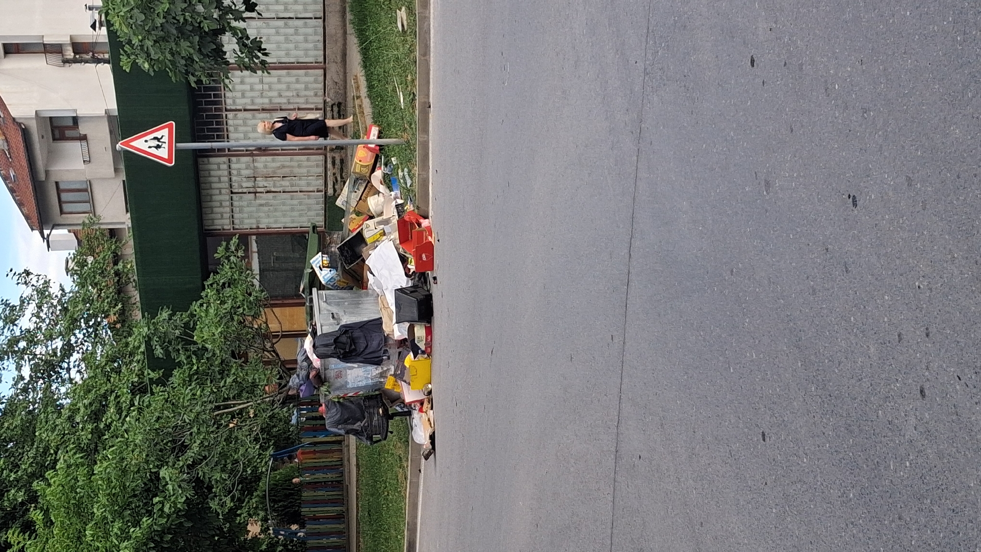 In Gotse Delchev, Bulgaria, a scene of overflowing trash bins and scattered refuse is depicted on a paved street adjacent to a grassy area and a building. A large, silver, commercial-style dumpster is situated near the curb, overwhelmed with garbage, some of which spills onto the pavement. Black trash bags and various discarded items, including cardboard boxes, plastic containers, and what appears to be broken furniture, are strewn around the dumpster and extend further onto the street.
To the right of the dumpster, a woman with blonde hair, wearing a black dress, walks away from the camera on the sidewalk. She is near a triangular road sign with a red border and a white interior, depicting two figures walking, indicating a pedestrian crossing. Behind the trash and the woman, a building with a dark green facade and large windows is visible. Trees with dense green foliage are present on the left and right sides of the frame, partially obscuring parts of the building and the sky. In the background, residential buildings with tiled roofs are also visible. The overall scene suggests a neglected public space with significant waste management issues.