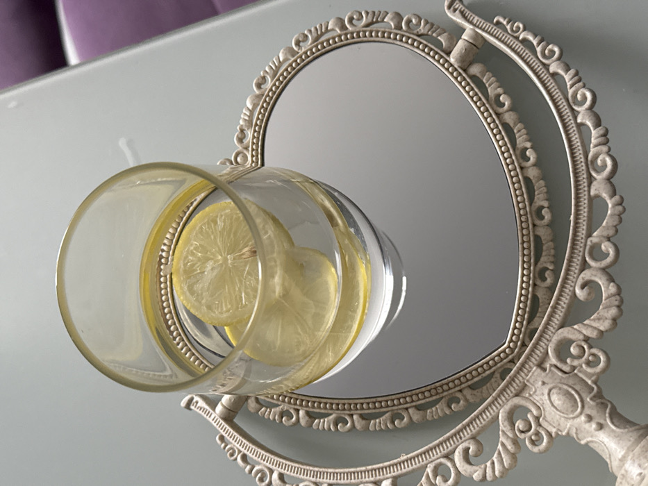 An overhead shot depicts a clear glass containing water and two yellow lemon slices, positioned centrally on a decorative, heart-shaped mirror with an ornate, light beige frame. The mirror features intricate scrollwork and a visible handle extending from its left side. Both the glass and mirror rest on a smooth, light gray tabletop, possibly made of glass. In the upper right background, a blurred segment of a purple upholstered surface is visible. There are no individuals present. The location is an indoor setting within Unknown, Unknown.