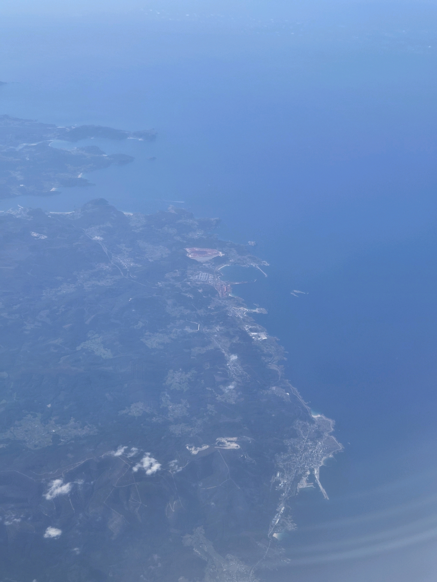 This is an aerial photograph taken from an airplane, looking down on a coastal area. The image displays a vast expanse of the ocean on the right side, meeting a rugged coastline on the left. The land is a mix of verdant hills, scattered buildings, and what appears to be industrial or port facilities near the coast. There are distinct inlets and peninsulas forming the coastline. In the foreground, clouds are visible, suggesting the airplane is flying above them or through them. The overall impression is one of a sparsely populated coastal region with evidence of human development, particularly along the waterfront. The specific location is identified as Barreiros, Spain.