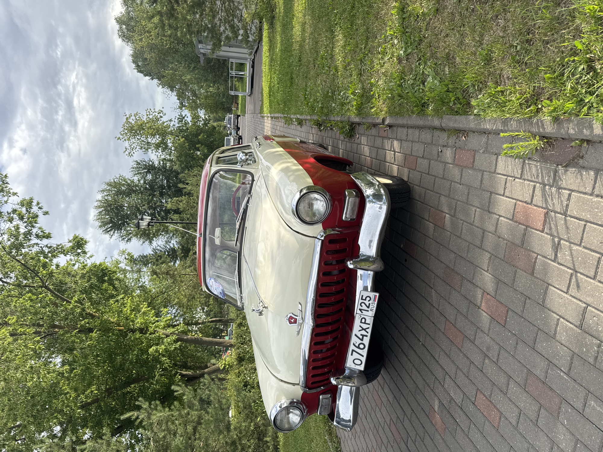 A two-tone red and cream vintage automobile is parked on a paved walkway. The car has chrome accents on the front grille, bumpers, and headlights. A license plate with Cyrillic script is visible on the front of the car. The car is surrounded by lush green trees and grass. The sky is overcast with scattered clouds. In the background, other vehicles and a lamppost are visible. The scene appears to be in a park or a public outdoor space.