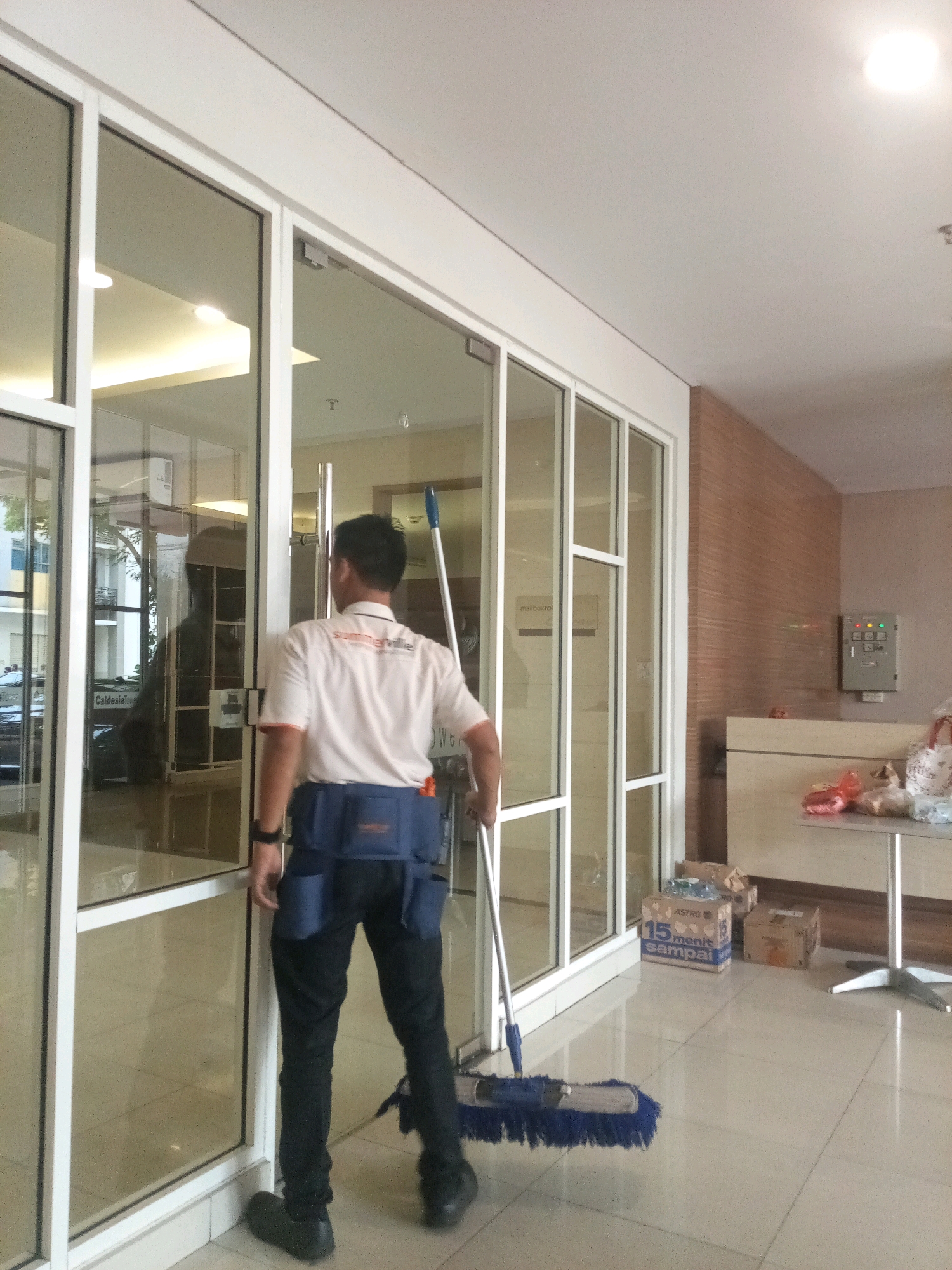 A male cleaner, wearing a white polo shirt with an orange collar and dark blue utility belt, is standing inside a building, facing a glass door. He is holding a mop with a blue brush head and a silver handle. The floor is tiled. The background shows a reception desk with items on it and a wall with a panel of buttons and lights. The scene appears to be an office building lobby in Bekasi, Indonesia, likely on the ground floor, given the view through the glass door which shows some outdoor elements.
