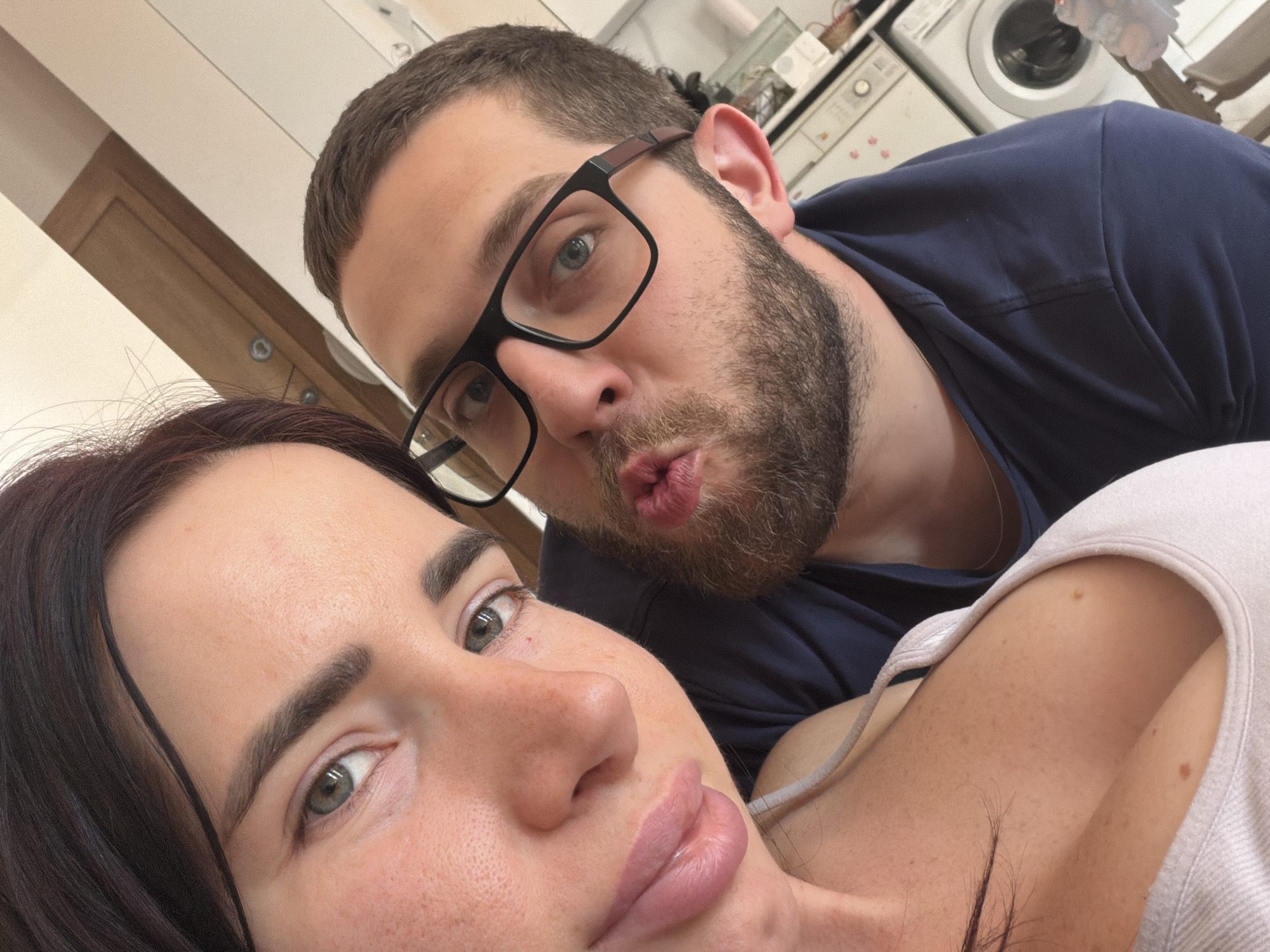 A close-up selfie captures a man and a woman in an indoor setting, likely a home in Sofia, Bulgaria. The woman occupies the foreground, her face partially visible on the left and bottom of the frame. She has dark hair, well-defined eyebrows, and light-colored eyes, looking towards the camera with a neutral expression. Her lips are full.
Behind and slightly above her, the man is visible on the right, looking over her shoulder. He has short brown hair, a beard, and is wearing black-framed glasses. He is playfully pursing his lips into a "kiss" shape and gazing directly at the camera with light-colored eyes. He is dressed in a dark blue t-shirt.
The blurred background reveals household appliances, including a washing machine and possibly other white goods, suggesting a utility area, kitchen, or laundry room. The image conveys an intimate and playful moment between the two individuals in a casual domestic environment. There is no visible text present in the image.