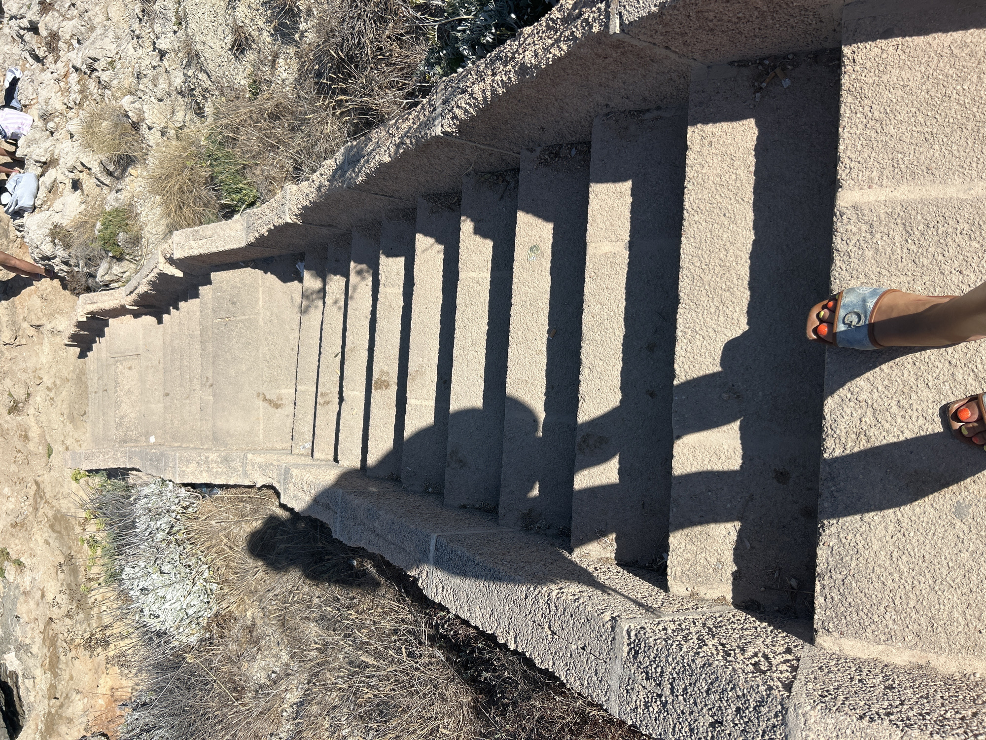 The image shows a person's feet wearing sandals as they stand on a concrete staircase. The staircase is outdoors, with dry vegetation and rocky terrain visible on either side. The shadows cast by the person and other unseen objects create a pattern on the stairs. In the background, there appears to be a beach or coastal area. The location is identified as Cala Lliteres, Spain.