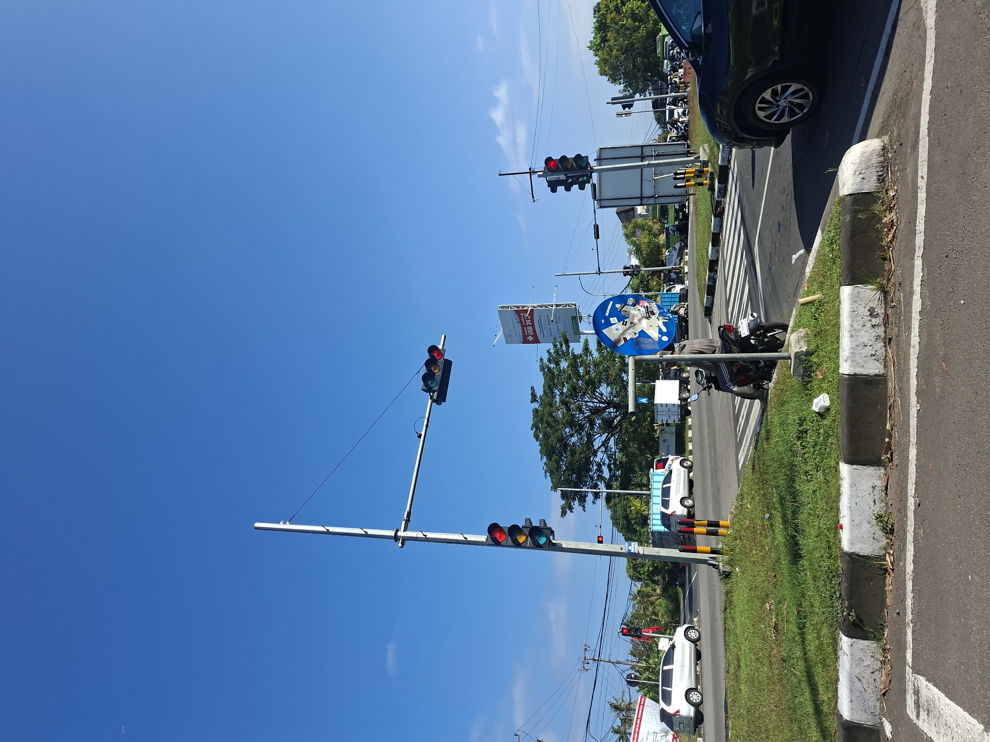 The image depicts a street intersection in Blahbatuh, Indonesia, under a clear blue sky. A traffic light pole with red and yellow lights illuminated is prominently featured in the foreground, along with a motorcycle parked on the side. Several vehicles, including cars and trucks, are visible on the road, some appearing to be stopped, possibly at the intersection. Green grass lines the roadside, and various signs and utility poles are scattered throughout the scene. The overall impression is of a busy urban environment on a sunny day.