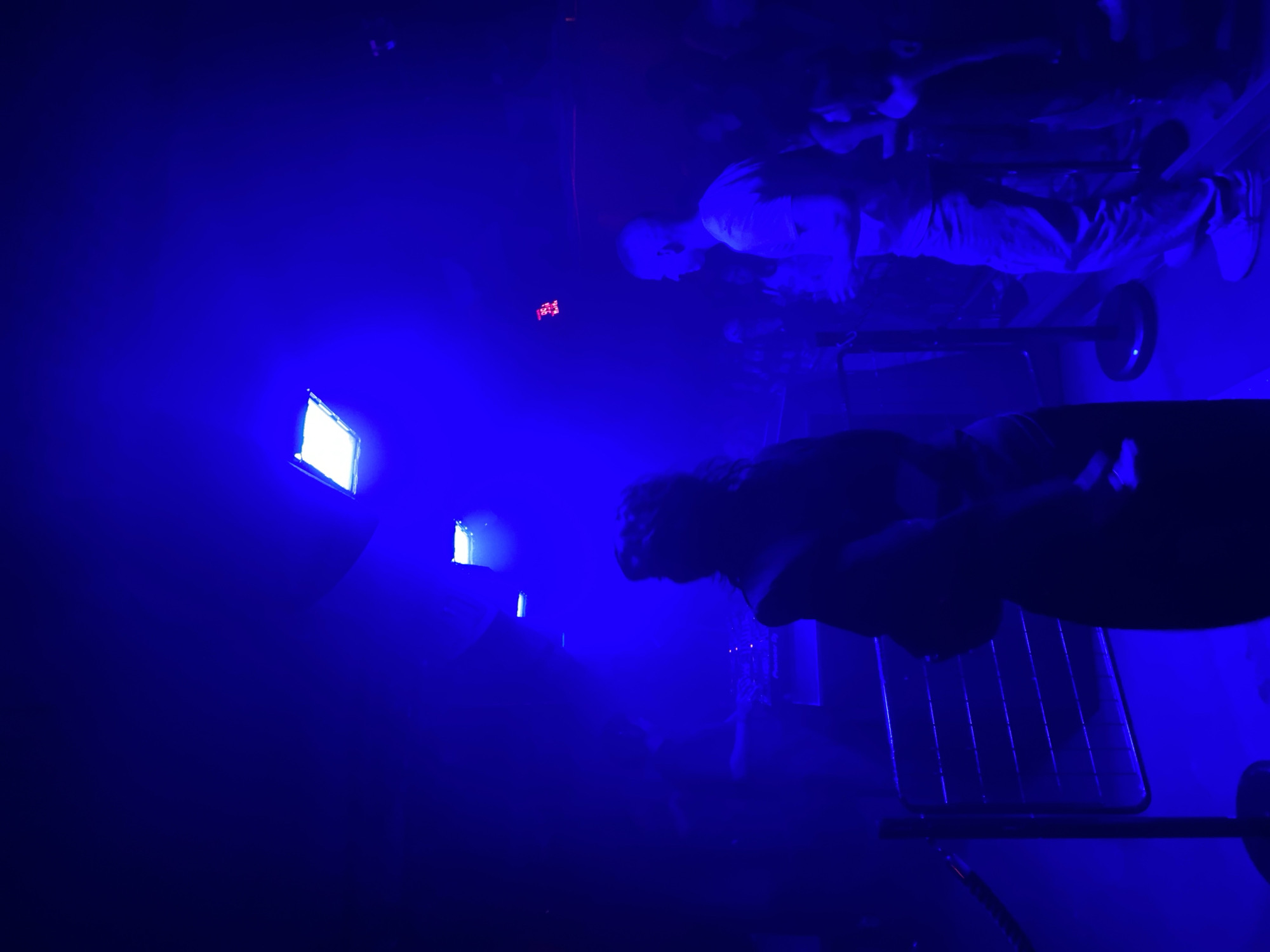 The image depicts an indoor scene within a nightclub or music venue in Paris, France, dominated by pervasive blue ambient lighting. Two rectangular light fixtures, emitting bright blue-white light, are visible on the ceiling. A red digital display, showing '35A Hz', is located in the upper right portion of the frame.
In the foreground, a heavily silhouetted individual with visible curly hair is positioned centrally, viewed from behind. To their right, in the midground, a male individual with a shaved head is seen in profile, facing left. He is wearing a light-colored t-shirt and trousers and appears to be holding a glass or bottle in his right hand.
On the left side of the frame, metal crowd control stanchions with horizontal bars are present, delineating a partitioned area. In the background, a DJ setup with associated equipment is visible, with another heavily silhouetted figure appearing to operate the console. Numerous other indistinct human figures populate the background and midground, forming a crowd. The intense blue lighting and potential atmospheric haze contribute to an overall low-visibility environment within the venue.