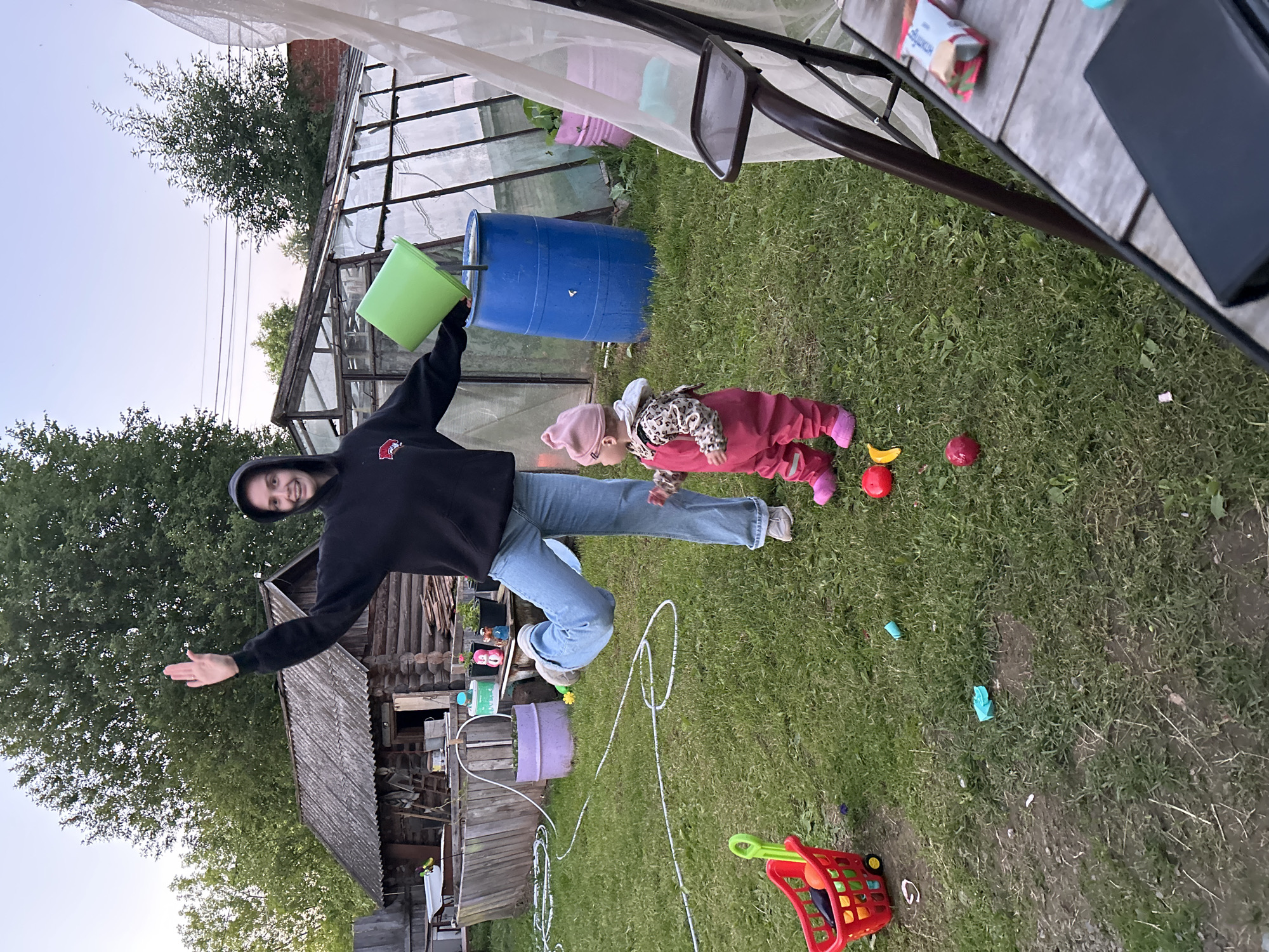 In the outdoor yard of a rural house in Азинский, Russia, a young woman stands with her right arm raised and a green bucket in her hand, appearing to be pouring its contents into a large blue barrel. She is wearing a black hooded sweatshirt and blue jeans. To her right, a toddler in a pink jumpsuit and hat stands looking forward. The yard is covered in green grass and scattered with children's toys, including a red toy shopping cart and several colorful balls. In the background, a rustic wooden house with a corrugated roof and a greenhouse with glass panels are visible. A white mosquito net is draped over a swing set on the right side of the frame, and a wooden table with a book and a snack wrapper is in the foreground. The overall scene depicts a casual, domestic setting.