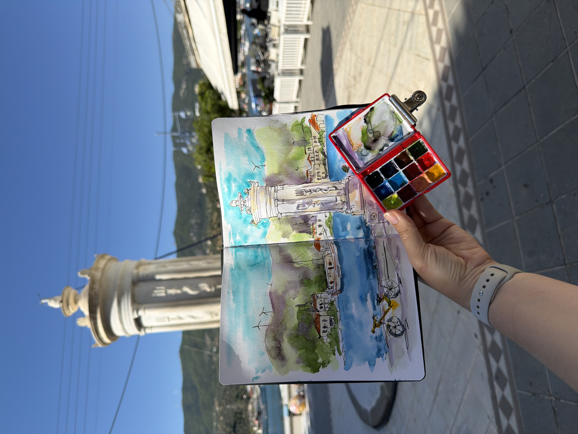 The image shows a person holding a sketchbook and watercolor palette in front of a view in Poros, Greece. The sketchbook displays a watercolor painting of the Poros Lighthouse, visible to the left. The palette is red with multiple colors of watercolor paint. The person is wearing a gray Apple Watch. The background includes the blue sky, mountains, trees, and buildings, suggesting a coastal town setting. The ground is paved with gray stone.