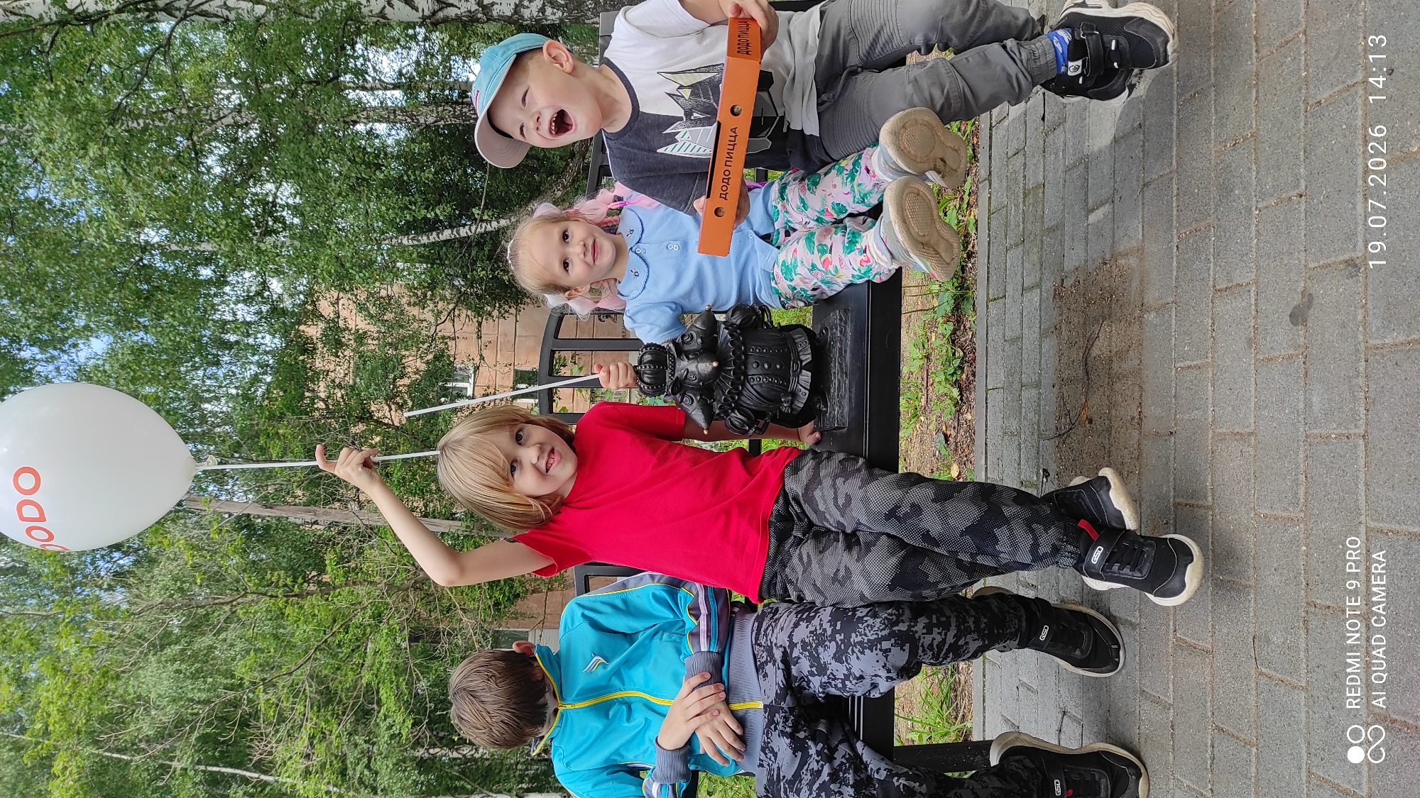 Four children are sitting on a park bench in a public park. The child on the far left is wearing a blue jacket and patterned pants. The child next to him is wearing a red t-shirt and patterned pants and holding a white balloon with "DODO" printed on it. The child in the middle is wearing a blue shirt and patterned pants. The child on the far right is wearing a t-shirt, gray pants, and a blue baseball cap, and is holding a pizza box. A small, dark statue of a creature is also on the bench between the two middle children. The background consists of trees and a building. The ground is paved with bricks. The timestamp "19.07.2026 14:13" is visible in the bottom right corner of the image. The location is identified as Chaikovsky, Russia.