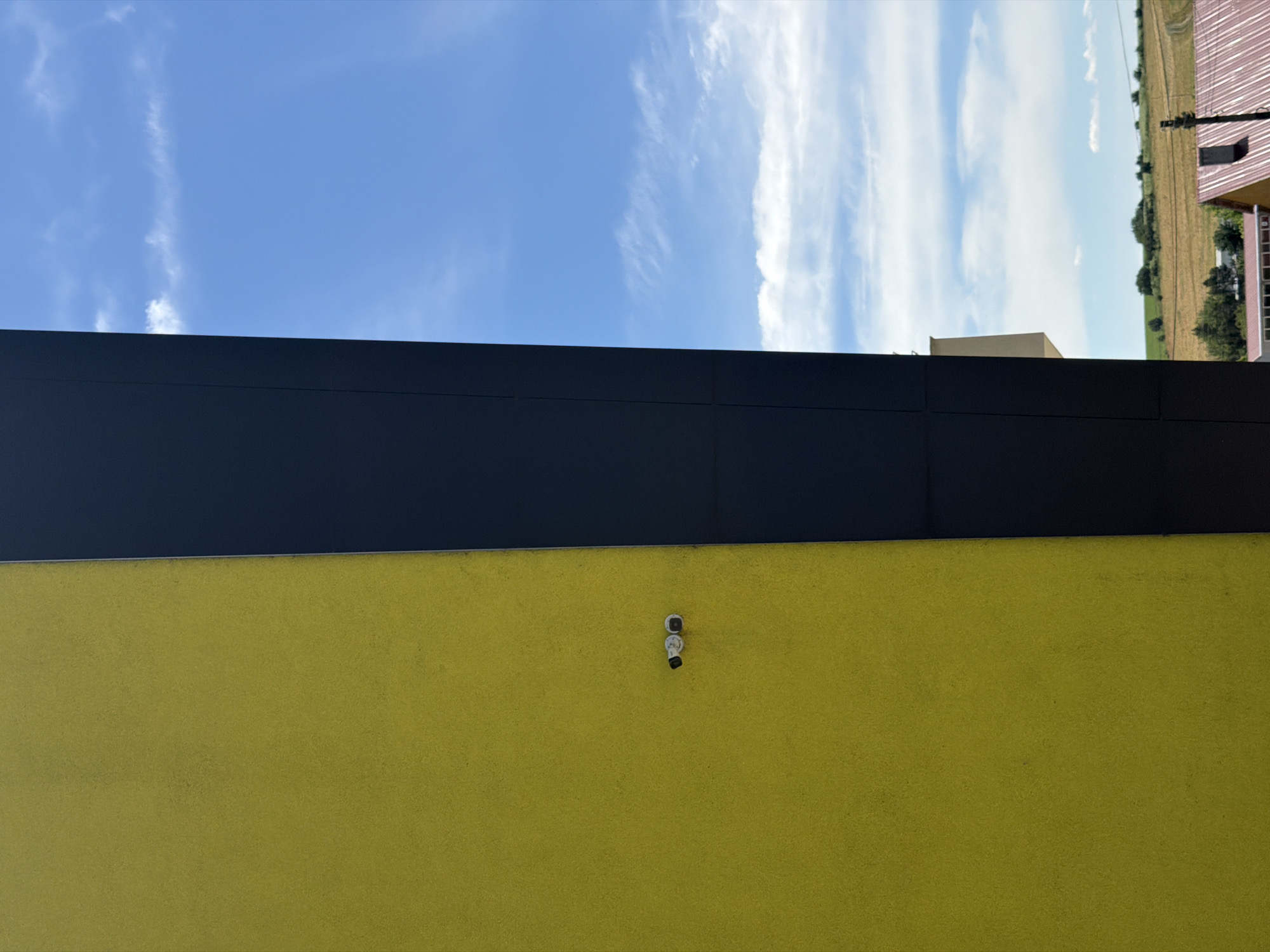 This image shows a section of a modern building with a bright yellow facade on the left and a dark gray facade on the right. Two small, white security cameras are mounted on the yellow wall. Beyond the building, a clear blue sky with scattered white clouds is visible, with hints of a rural landscape, including fields and some rooftops, in the distance. The scene is captured from a slightly elevated perspective, looking upwards. The location is identified as Dolno Ablanovo, Bulgaria, suggesting this is an exterior view of a building in that area.