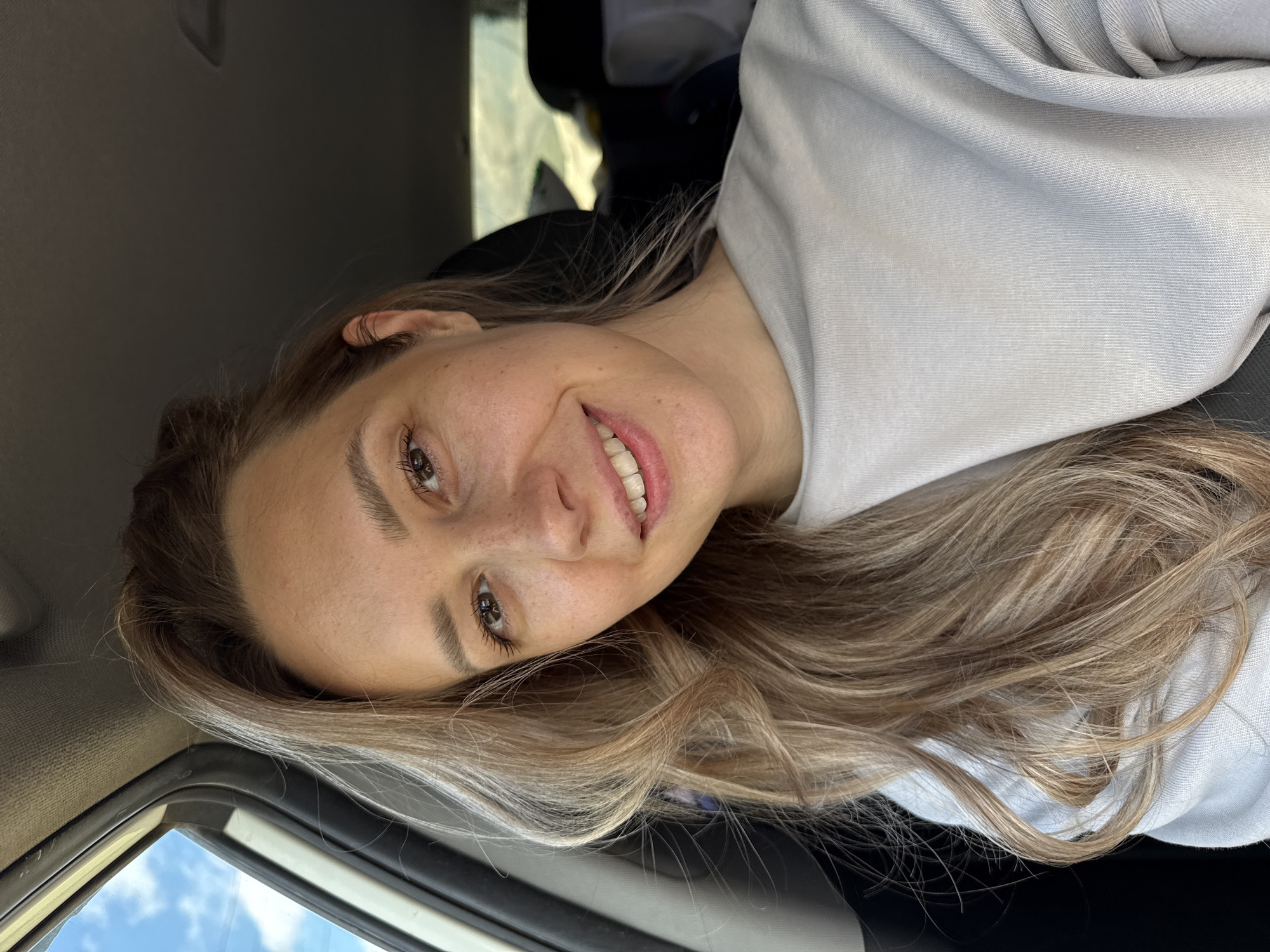 A young Caucasian woman with blonde hair is pictured from the chest up, looking directly at the camera. She is seated in the passenger seat of a car, with the car's interior visible in the background. The woman is wearing a light gray crewneck sweatshirt. Her right side is framed by the car's window, through which a blue sky with some clouds can be seen. The overall lighting suggests it is daytime.