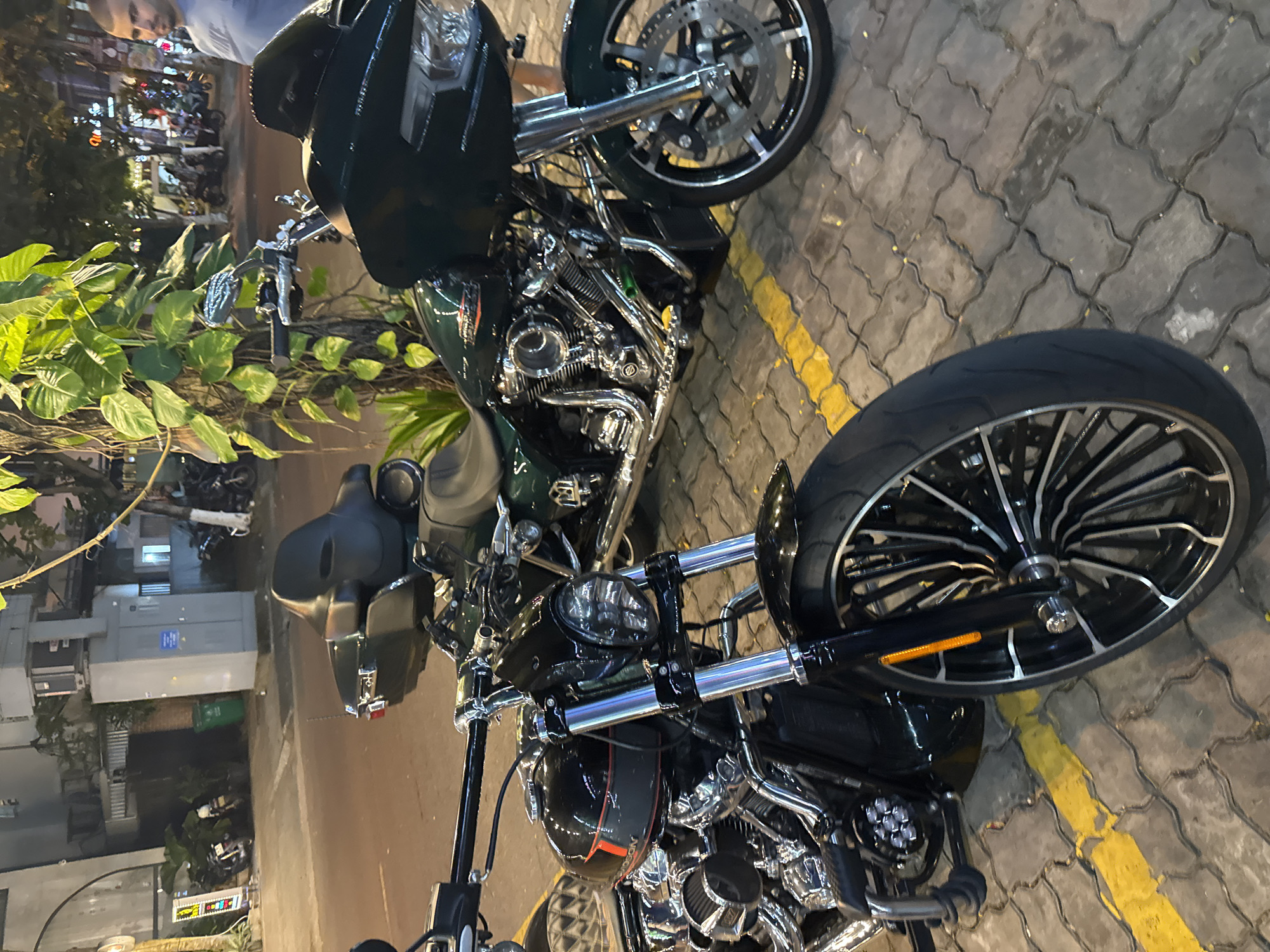 Two motorcycles are parked on a brick walkway next to a street. One motorcycle is green, while the other is black. The street is lined with trees and buildings. Other people can be seen in the background. Đà Nẵng, Vietnam.
